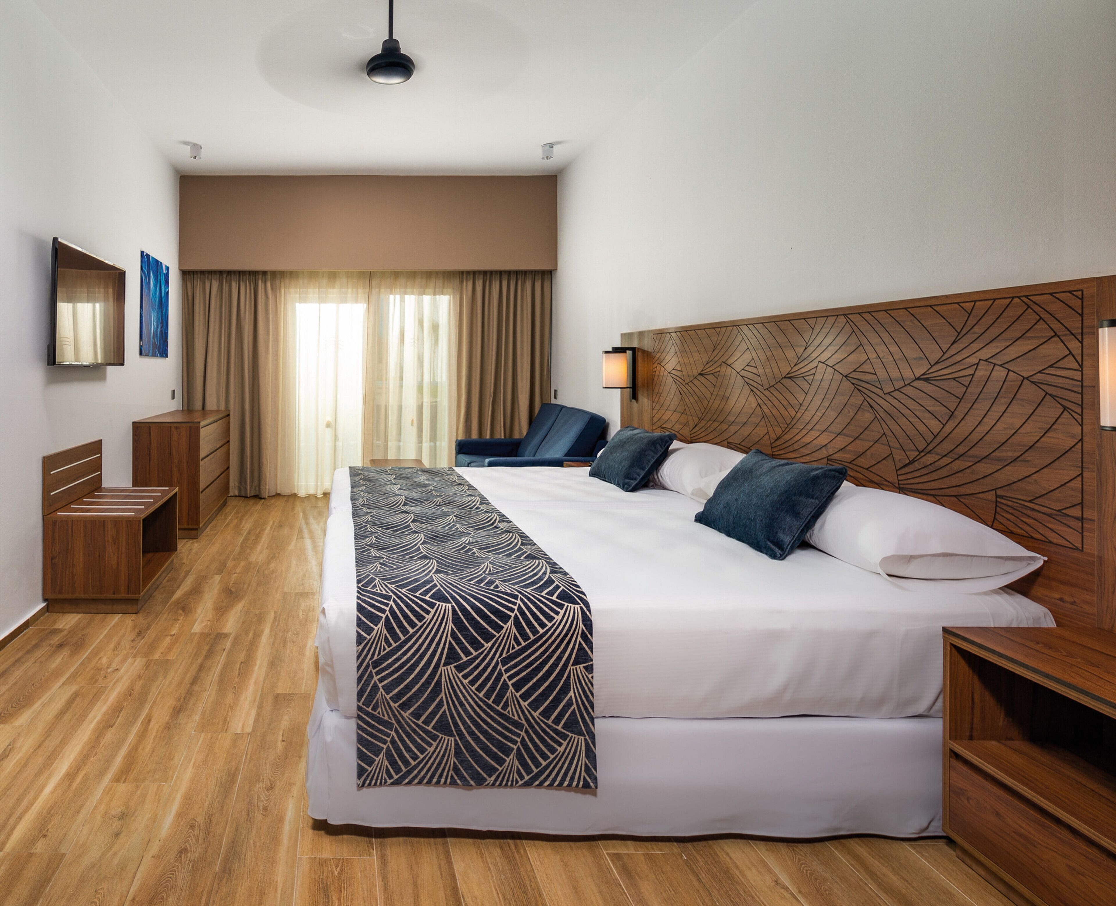 Une chambre d’hôtel avec un grand lit, une tête de lit en bois, un bureau, une chaise, une armoire et un miroir.