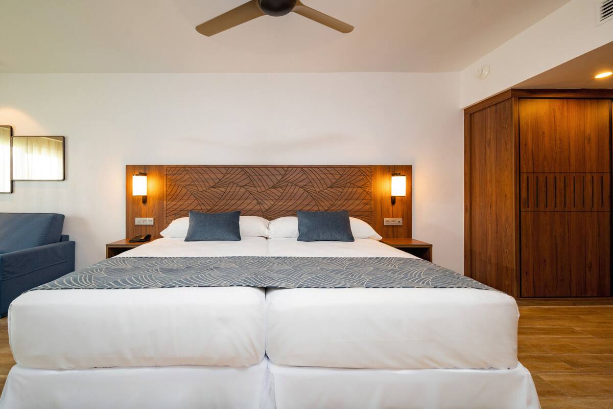 Habitación de hotel moderna con una cama grande, dos mesitas de noche con lámparas, un ventilador de techo, un sillón azul y un armario de madera.