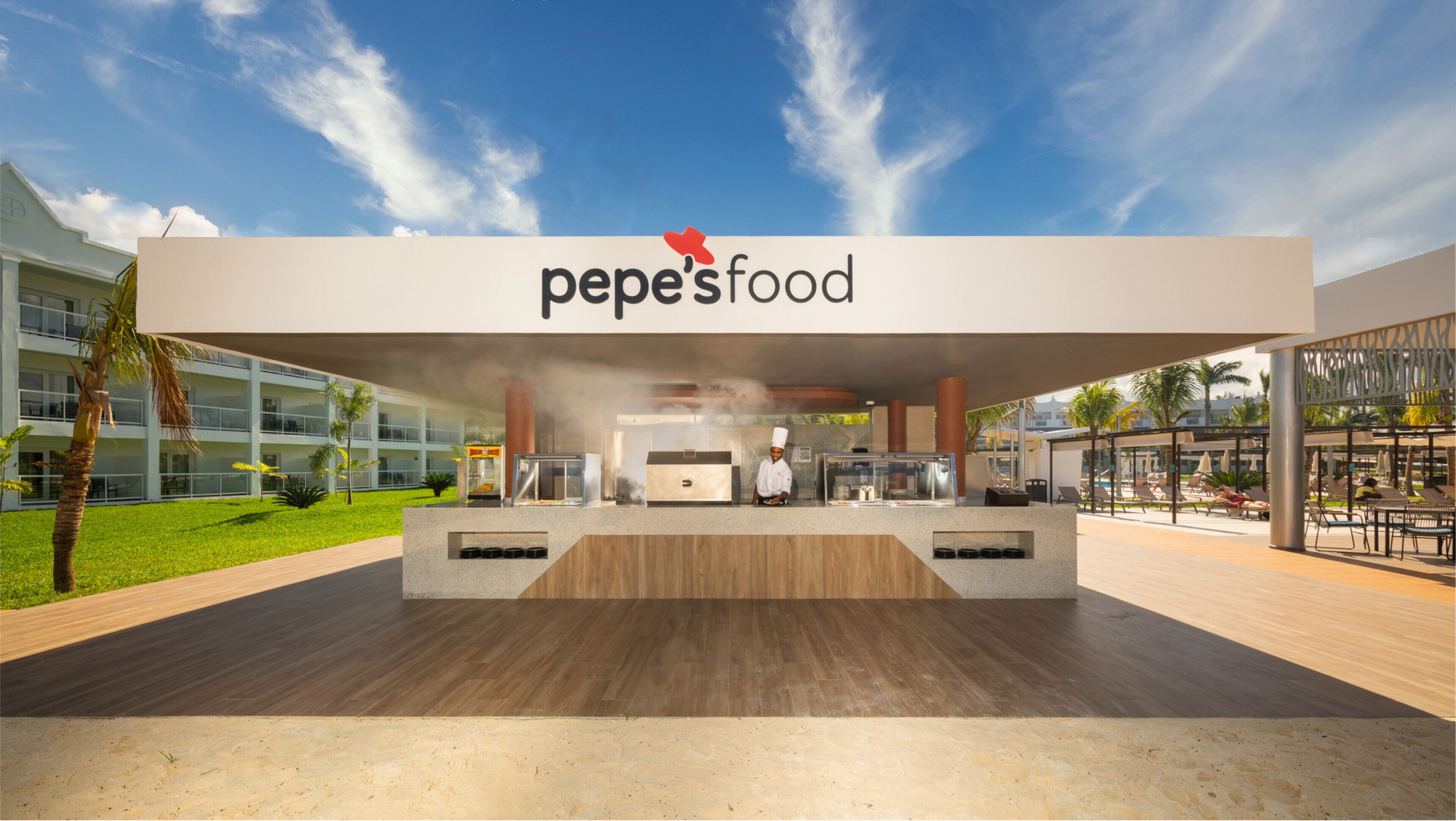 Zona pranzo all'aperto da Pepe's Food, con uno chef che prepara piatti, circondata da un edificio moderno e una zona piscina.