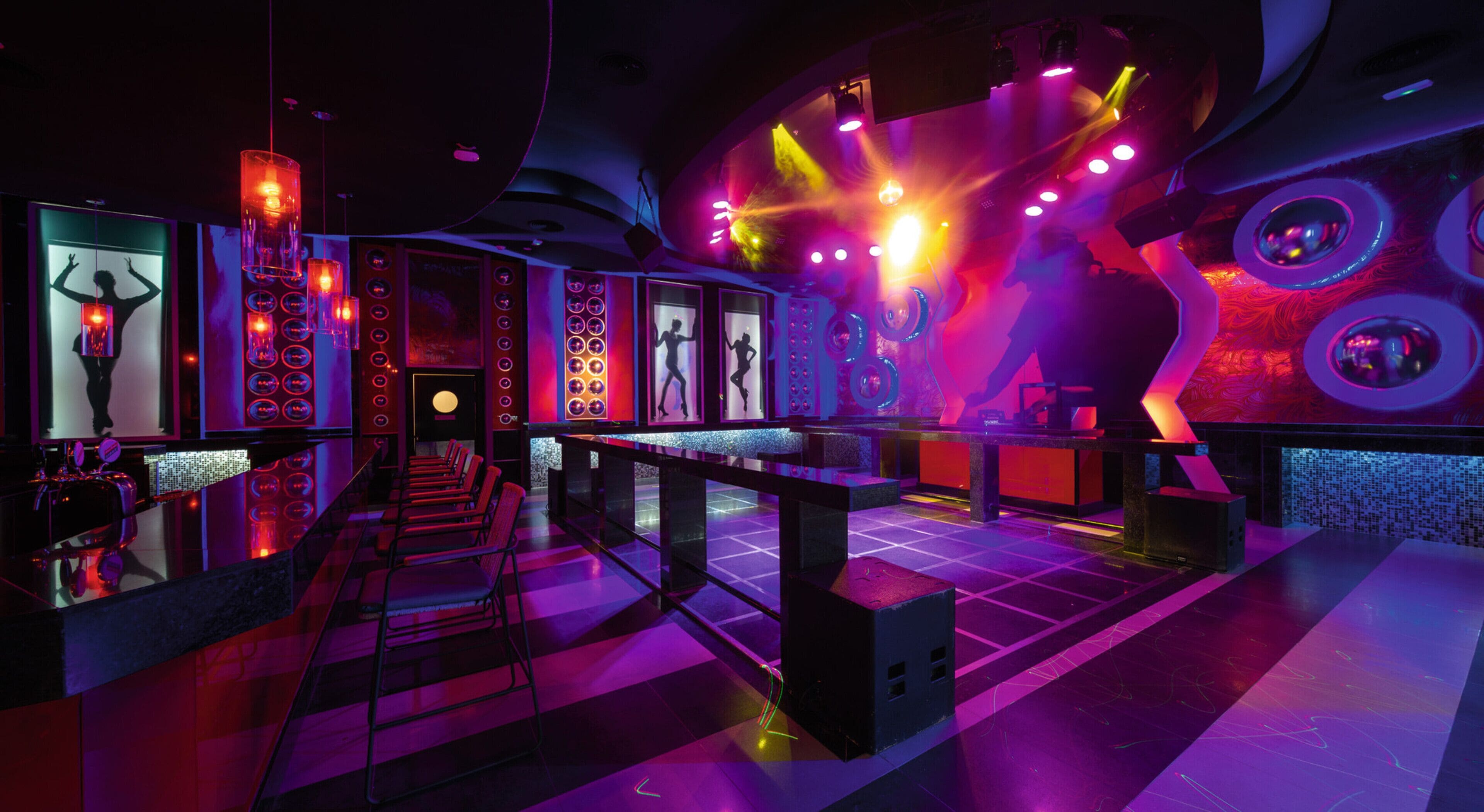 A modern bar with a long counter, multiple seating areas, and vibrant lighting.