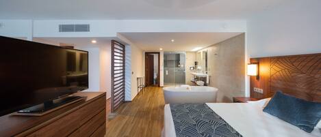 Ein modernes Hotelzimmer mit Flachbildfernseher, Holzkommode, Bett mit gemusterten Betttextilien, einem großen Fenster und einem Badezimmer mit Badewanne und Spiegel.