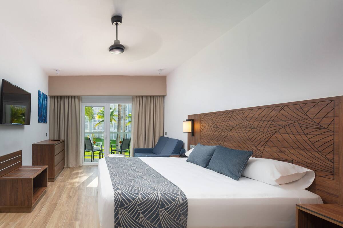 Una habitación de hotel moderna con una cama grande, cabecera de madera, televisión de pantalla plana, escritorio y balcón con vistas a palmeras.