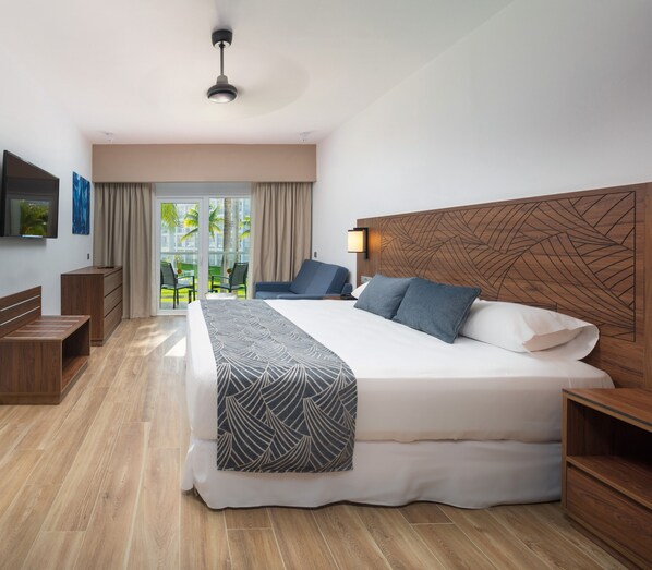 Una habitació d'hotel moderna amb un llit gran, un reposacapses de fusta, una televisió de pantalla plana, un escriptori i un balcó amb vista a palmeres.