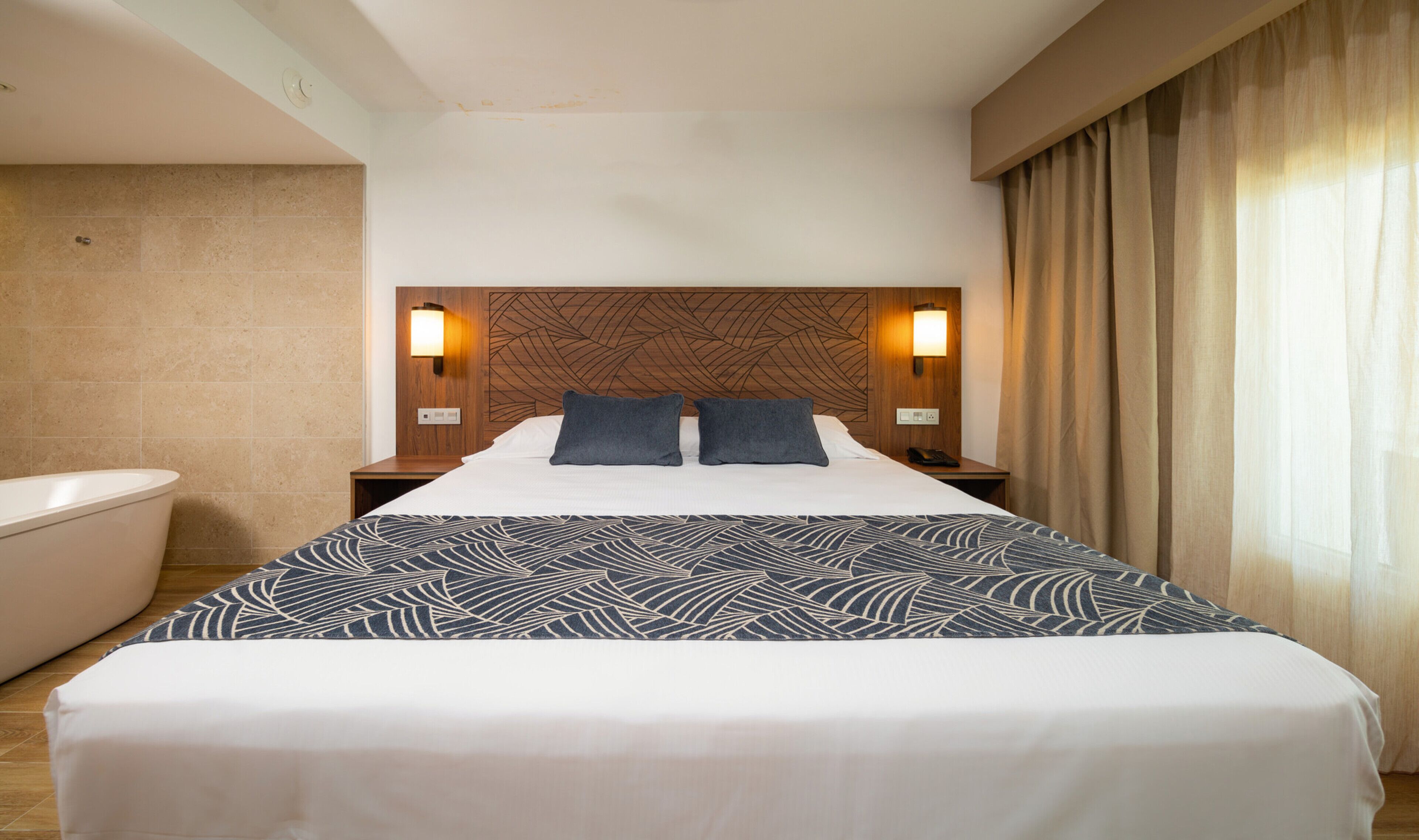A hotel room with a large bed, two bedside lamps, a headboard with a geometric pattern, a bathtub, and a window with curtains.