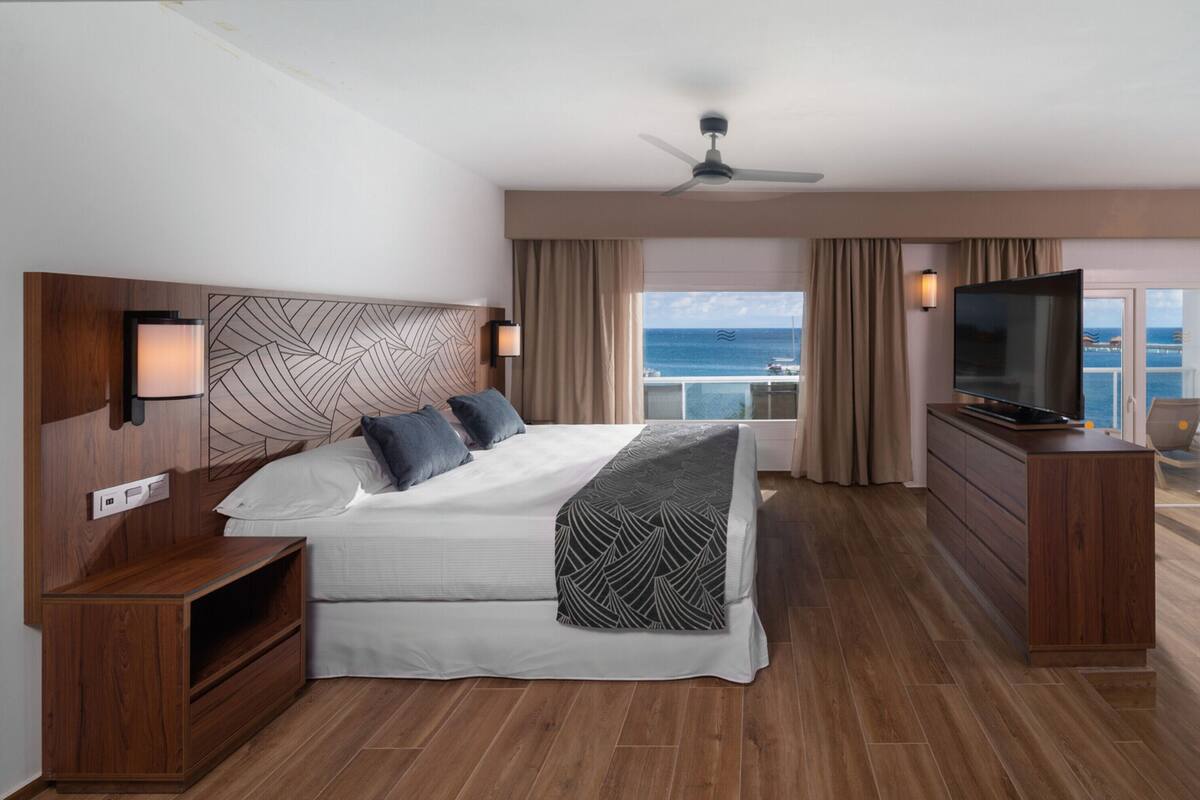 Habitación de hotel moderna con una cama grande, televisor, escritorio y balcón con vista al mar.
