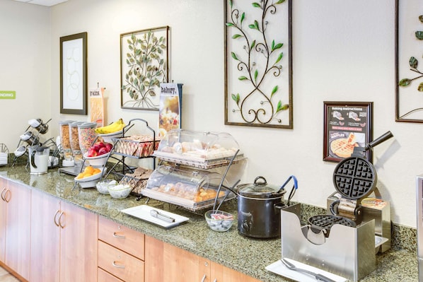 A buffet counter with various food items, including fruits, pastries, and a waffle maker. - Microtel Inn & Suites by Wyndham Saraland/North Mobile (Saraland)