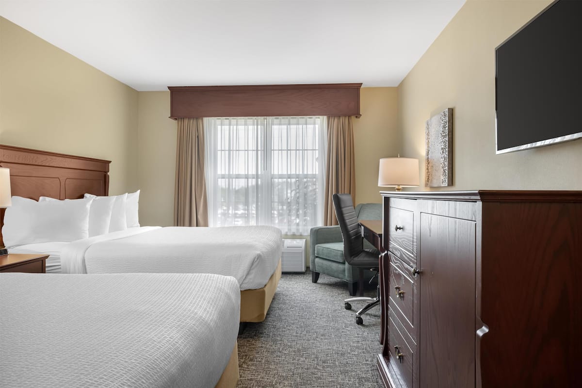 Une chambre d’hôtel avec deux lits, un bureau, une chaise, une armoire et une grande fenêtre avec des rideaux.