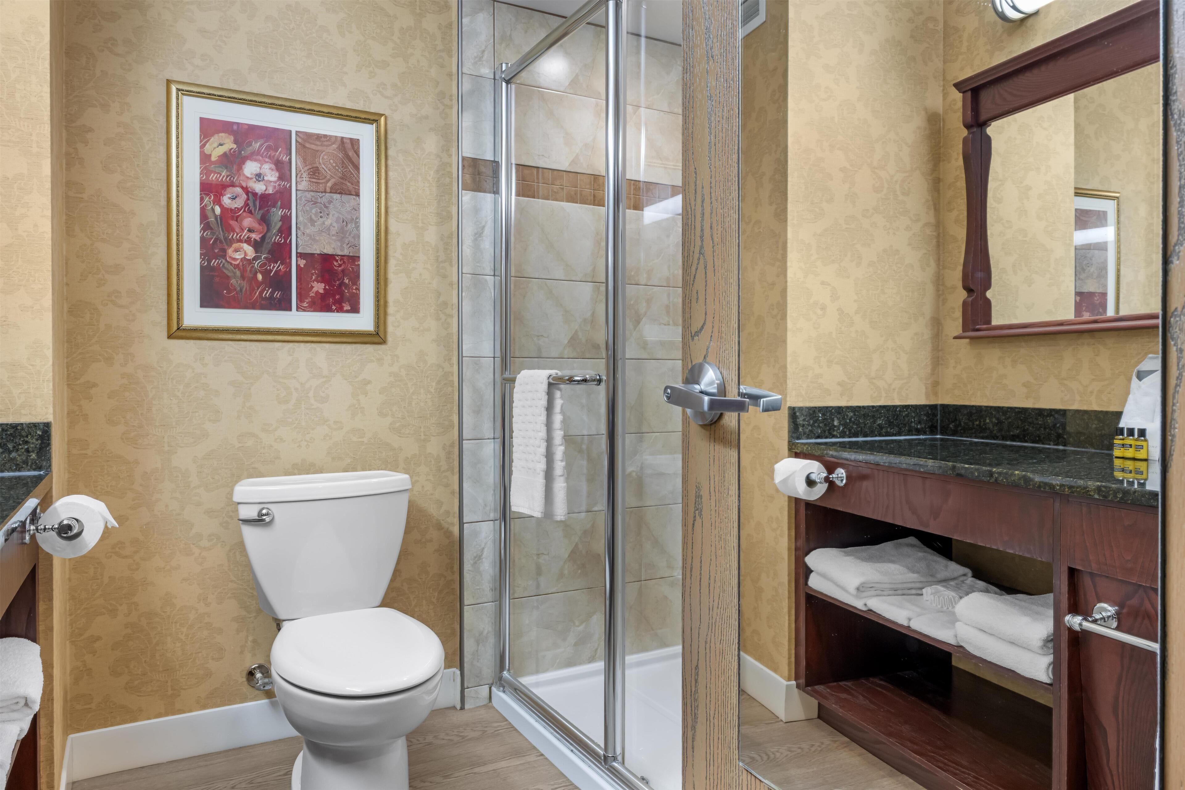 Un baño con inodoro, cabina de ducha de vidrio, tocador de madera con espejo y una obra de arte floral enmarcada en la pared.
