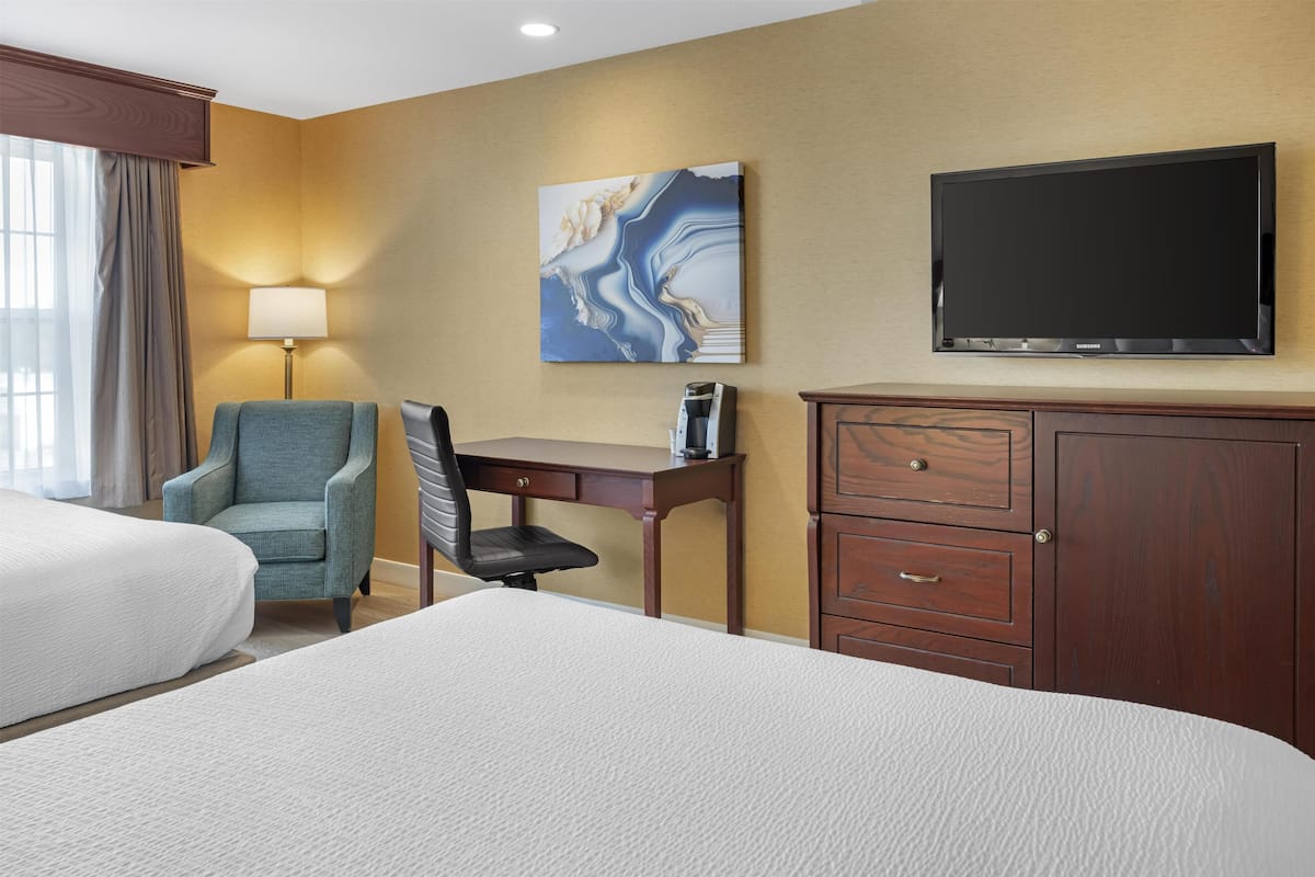 Une chambre d’hôtel avec deux lits, un bureau avec une chaise, un téléviseur à écran plat, un tableau au mur et une armoire en bois.