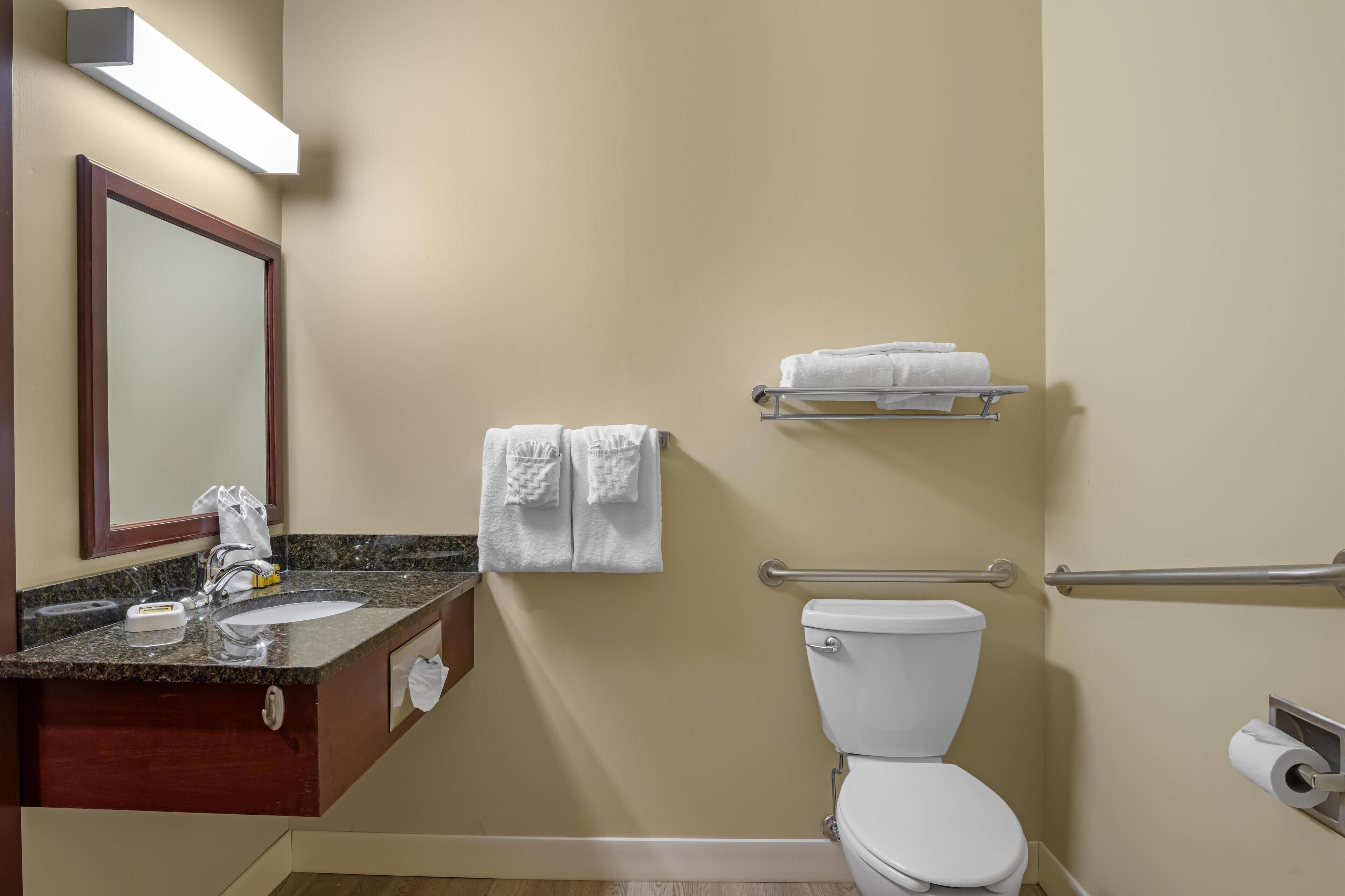 Un baño con lavamanos, espejo, toallero, inodoro y barras de apoyo.