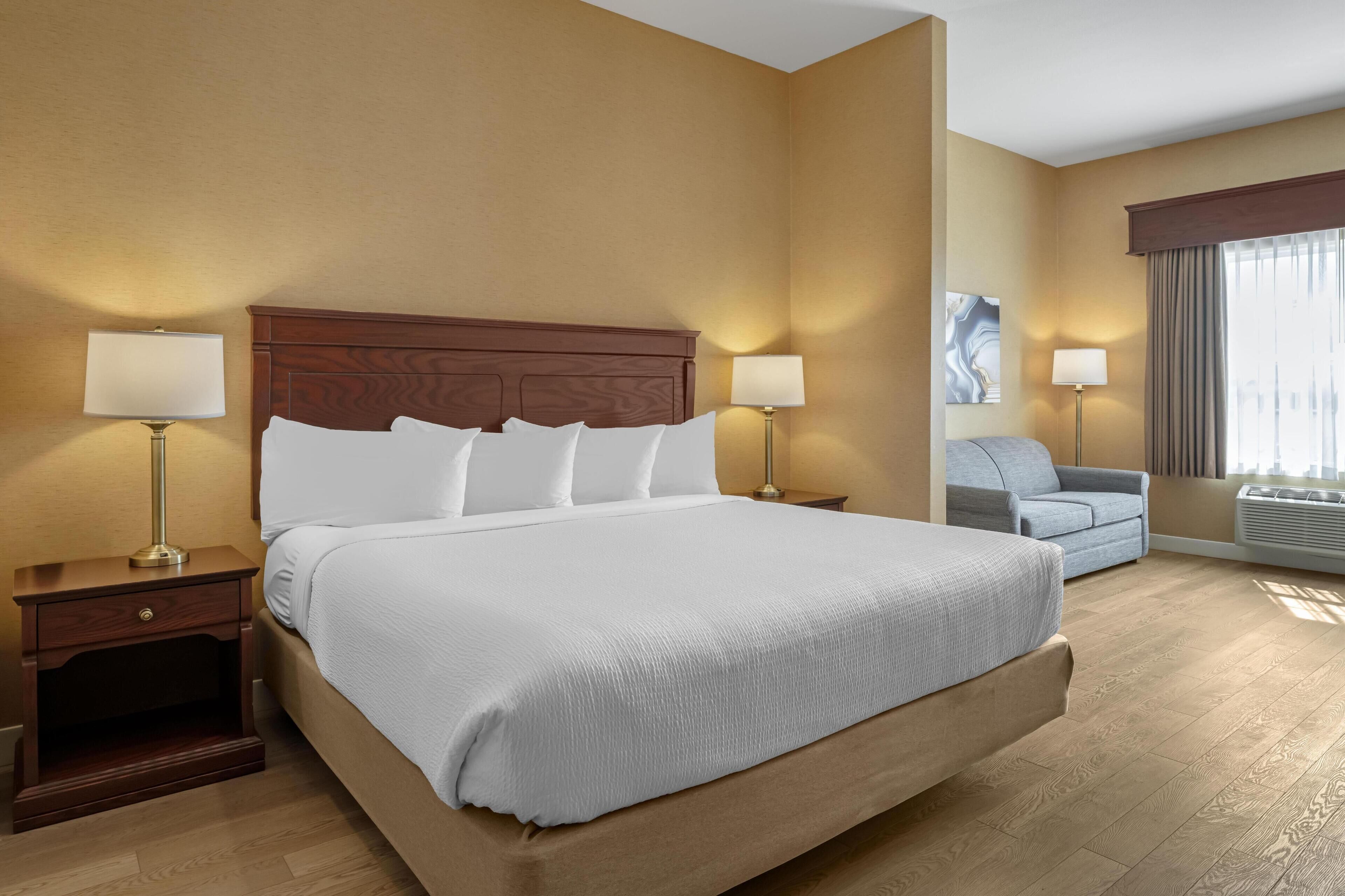A hotel room with a large bed, two bedside tables with lamps, a grey sofa, and a large window with curtains.