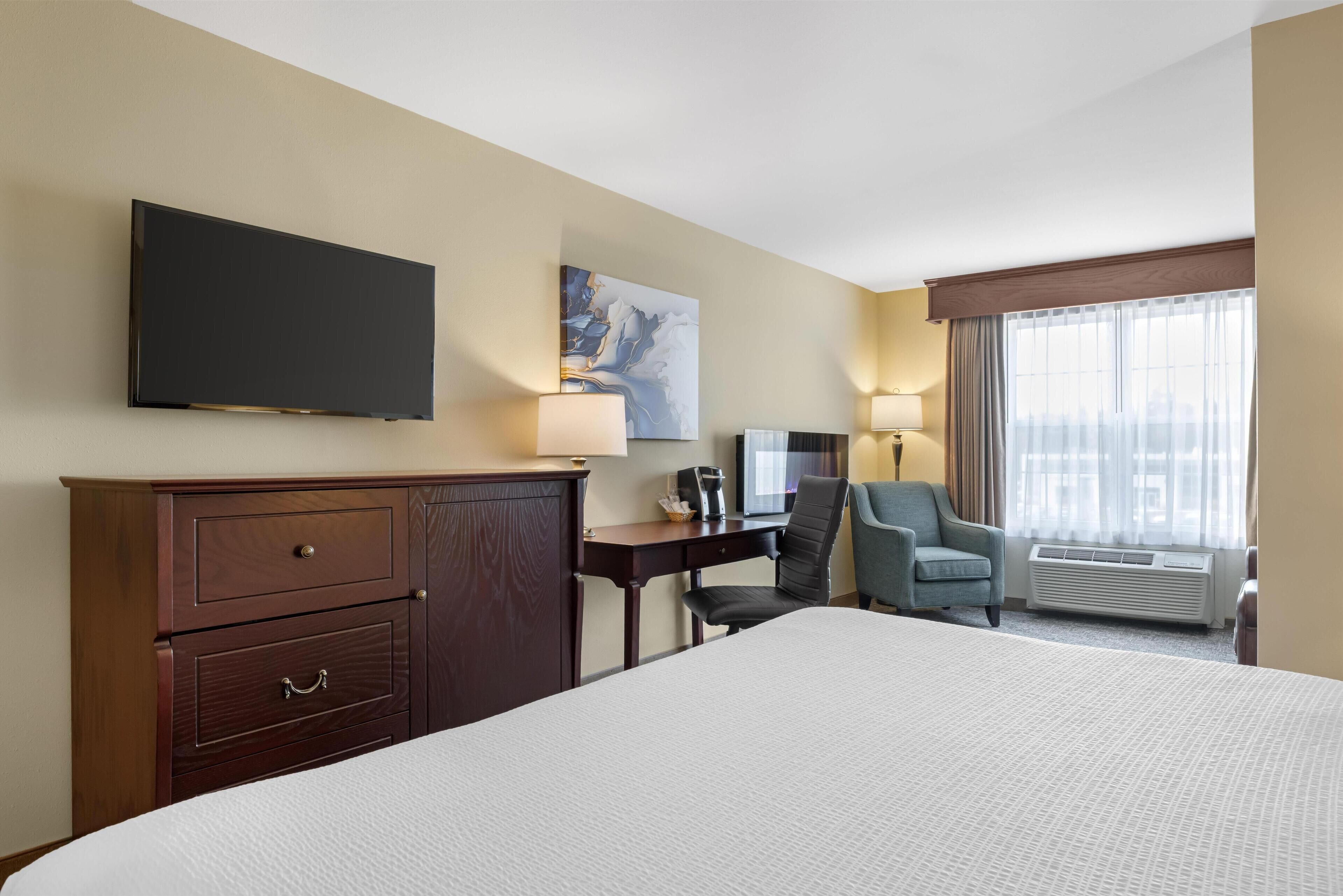 A hotel room with a large bed, a desk with a chair, a TV mounted on the wall, a painting, and a window with curtains.