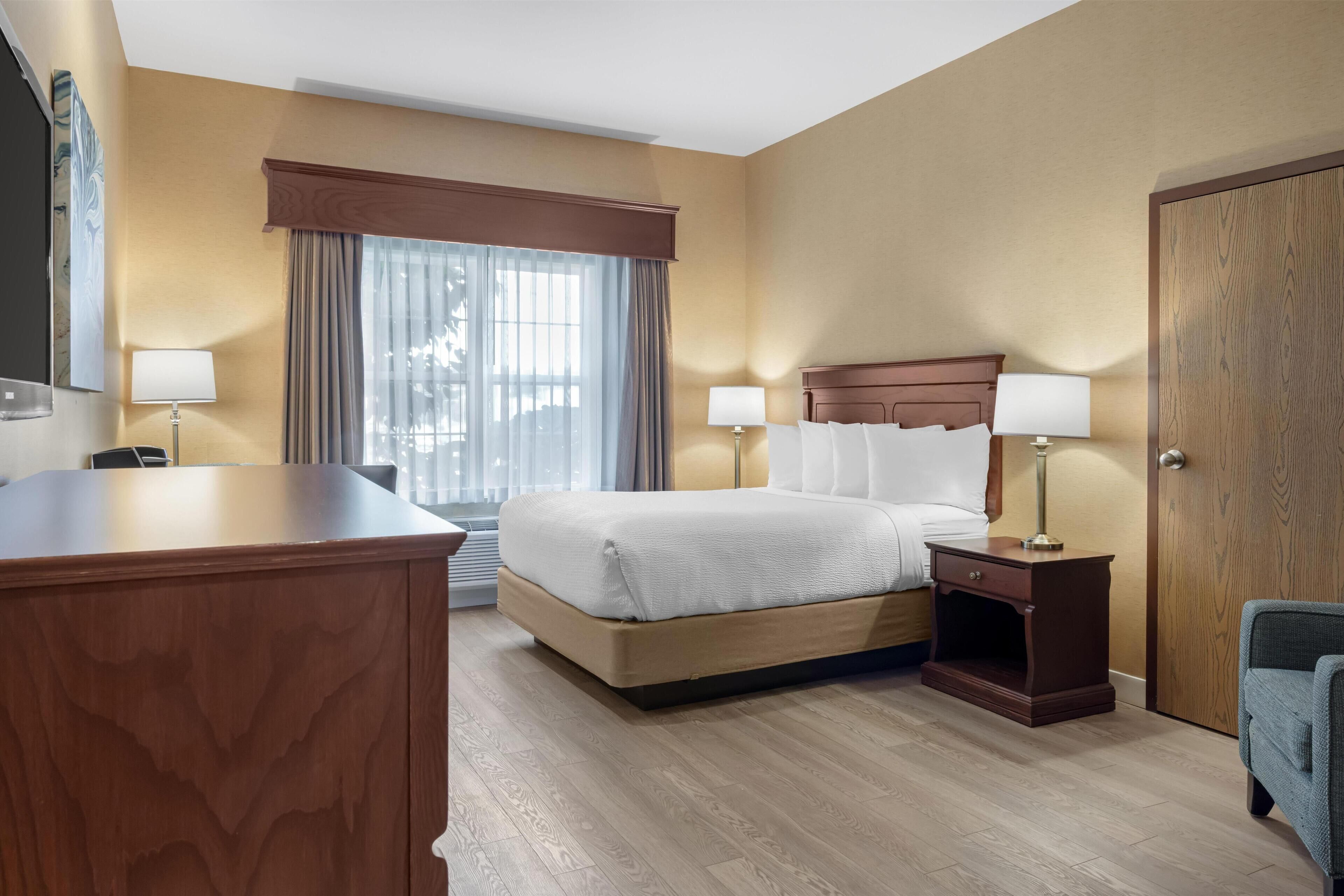 Quarto de hotel com uma cama grande, duas mesas de cabeceira com luminárias, uma escrivaninha de madeira e uma porta.