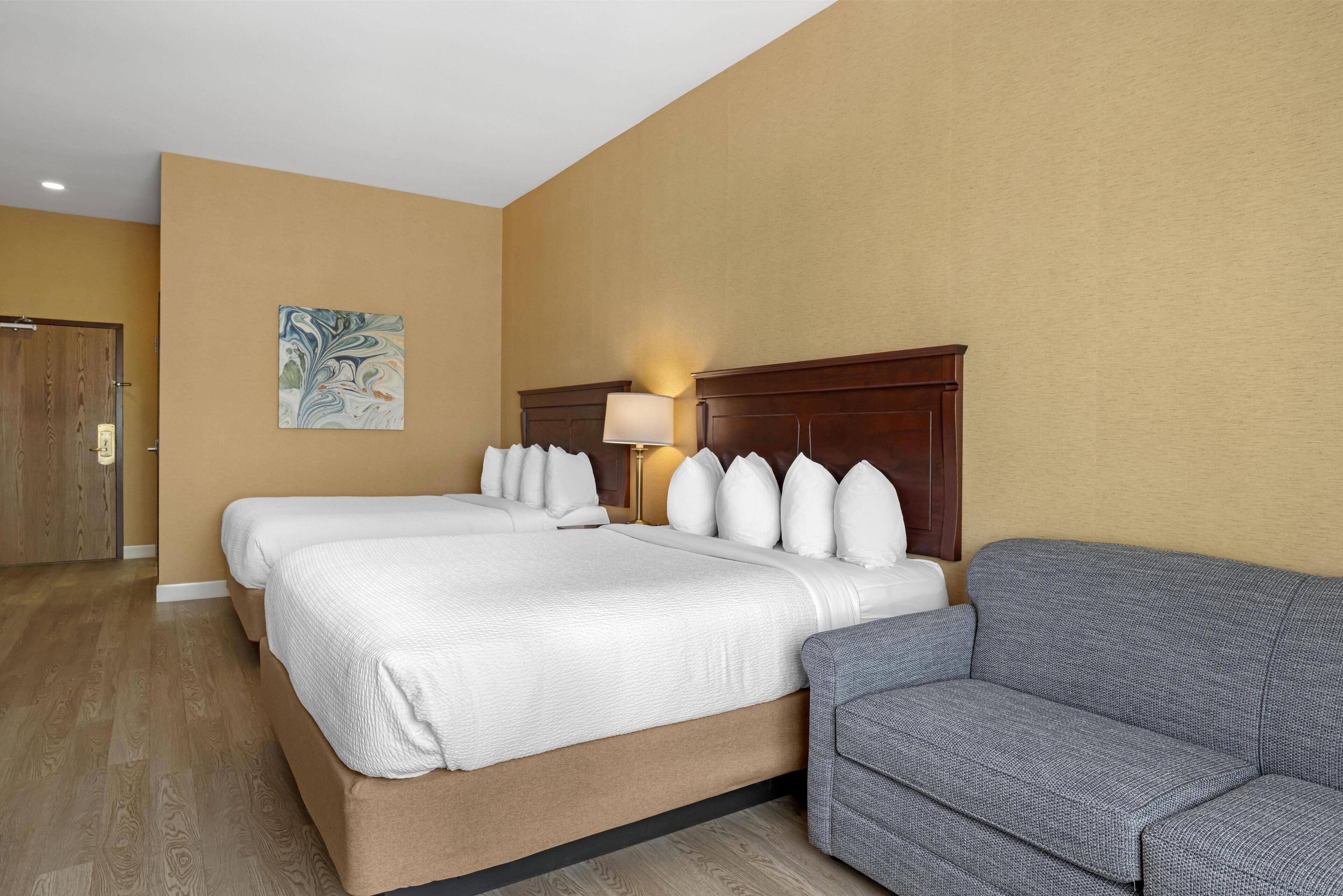 A hotel room with two beds, a gray sofa, a wooden door, and a wall painting.