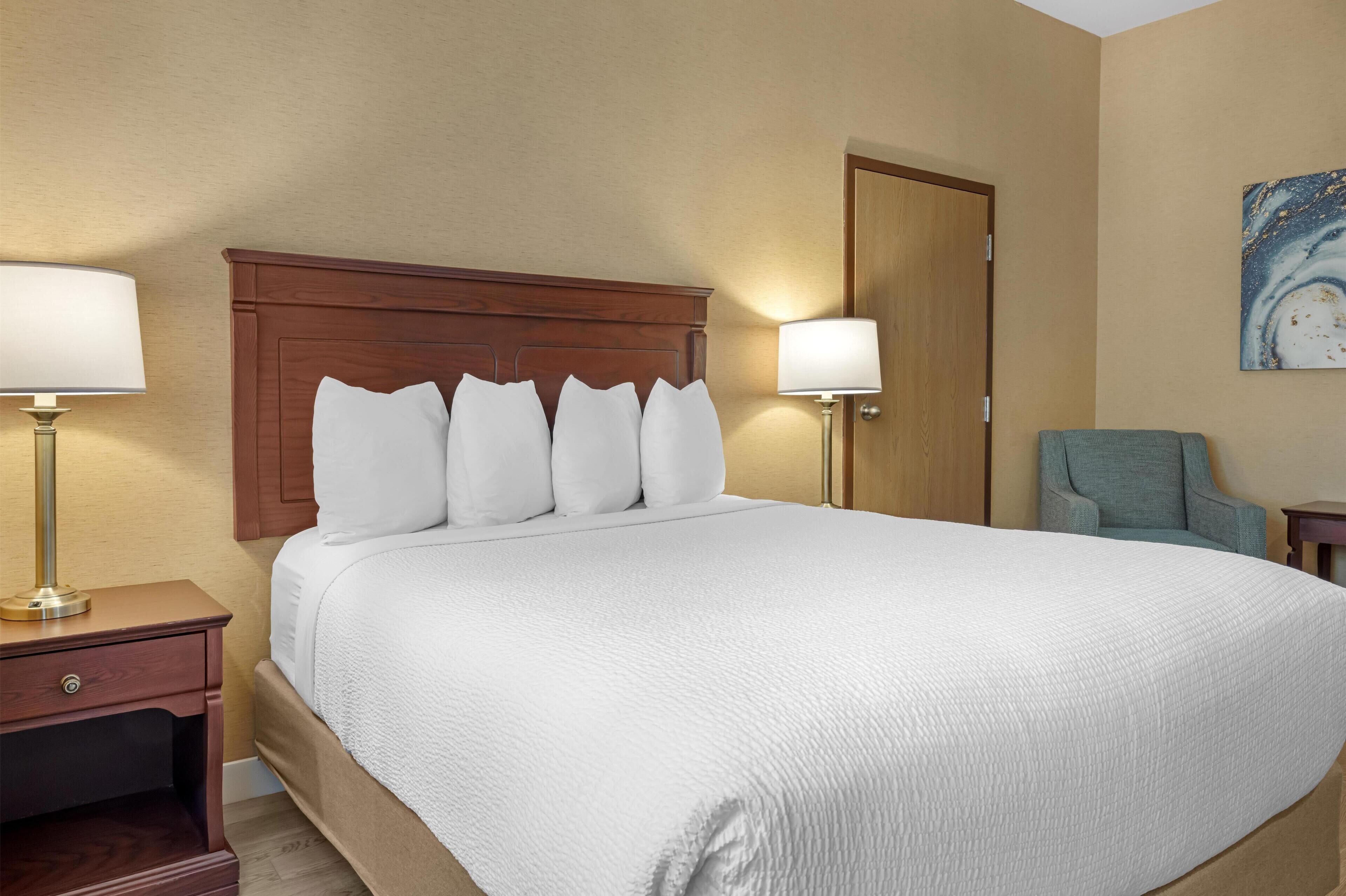 Une chambre d’hôtel avec un grand lit, deux lampes de chevet, une tête de lit en bois, une porte et une chaise.