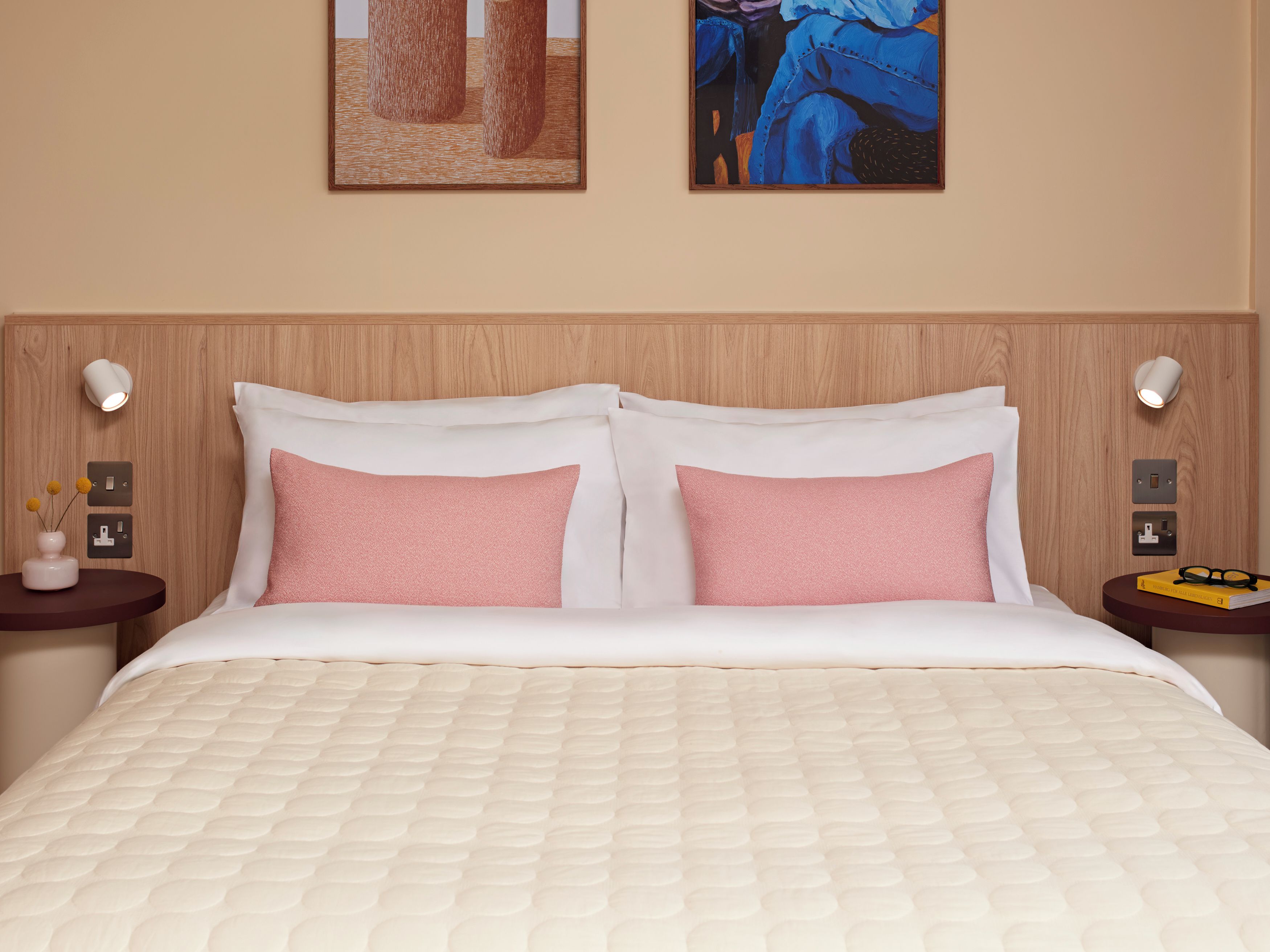 A hotel room with a wooden headboard, two bedside tables with lamps, a bed with white bedding and pink pillows, and artwork on the walls.