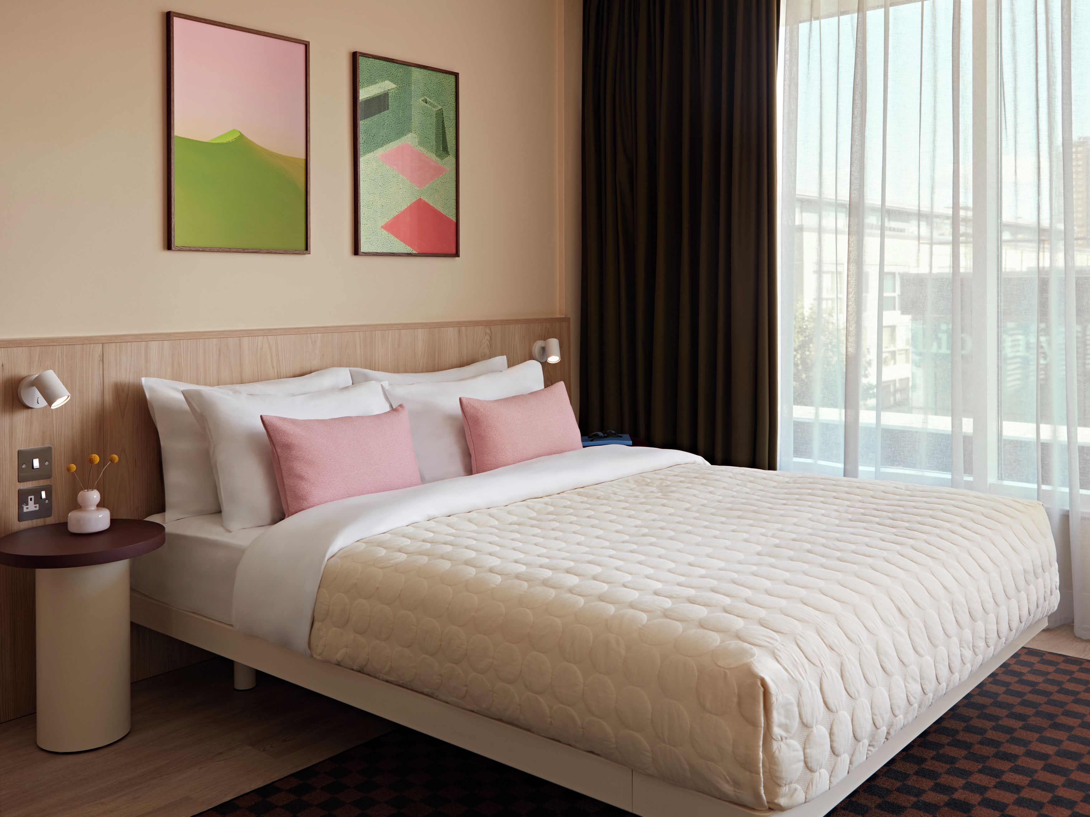 Une chambre d’hôtel avec un lit, une table de chevet, des œuvres d’art mural et une fenêtre avec des rideaux.