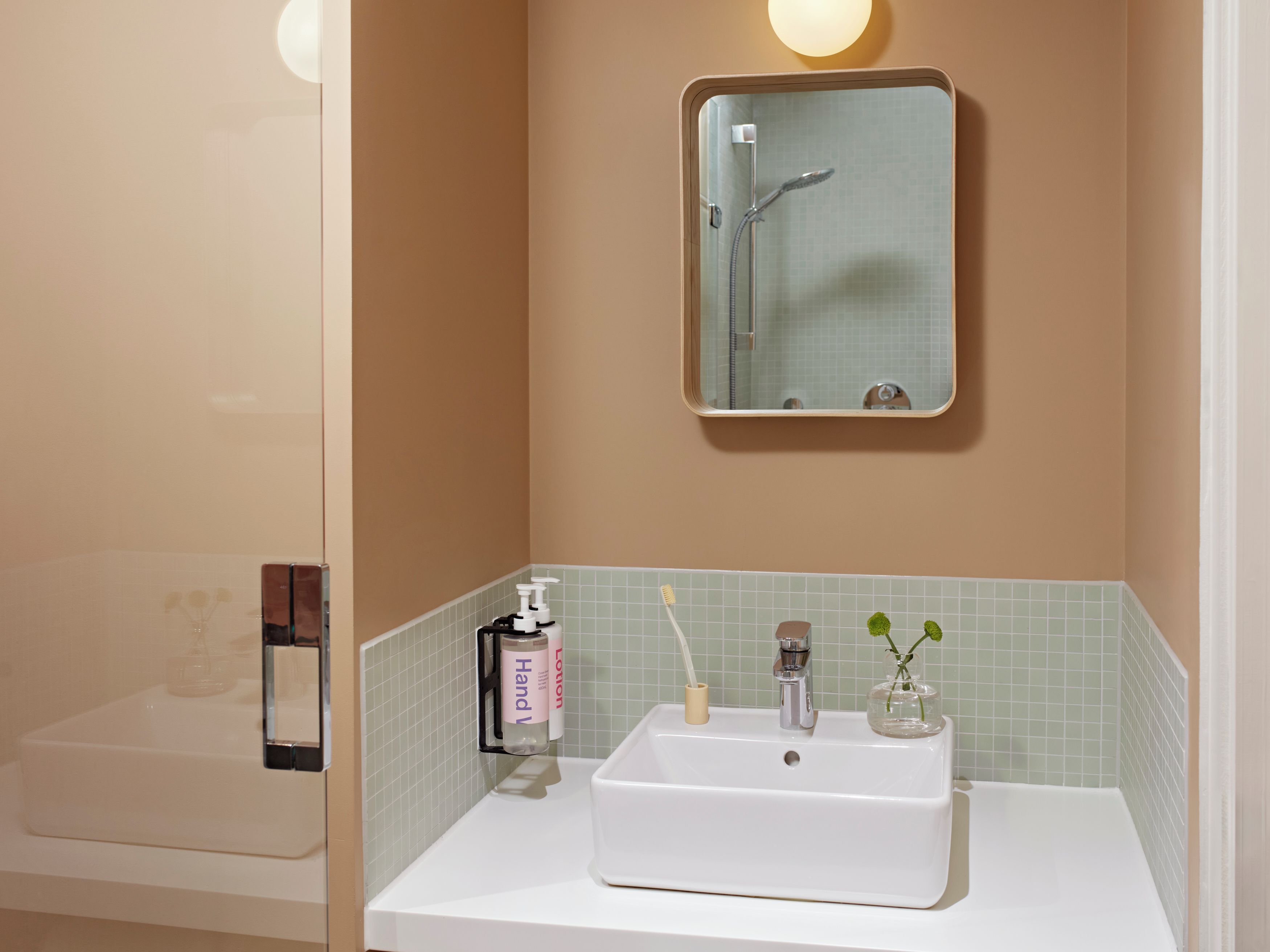 A bathroom with a white sink, a mirror, a handheld showerhead, and a small vase with a plant.