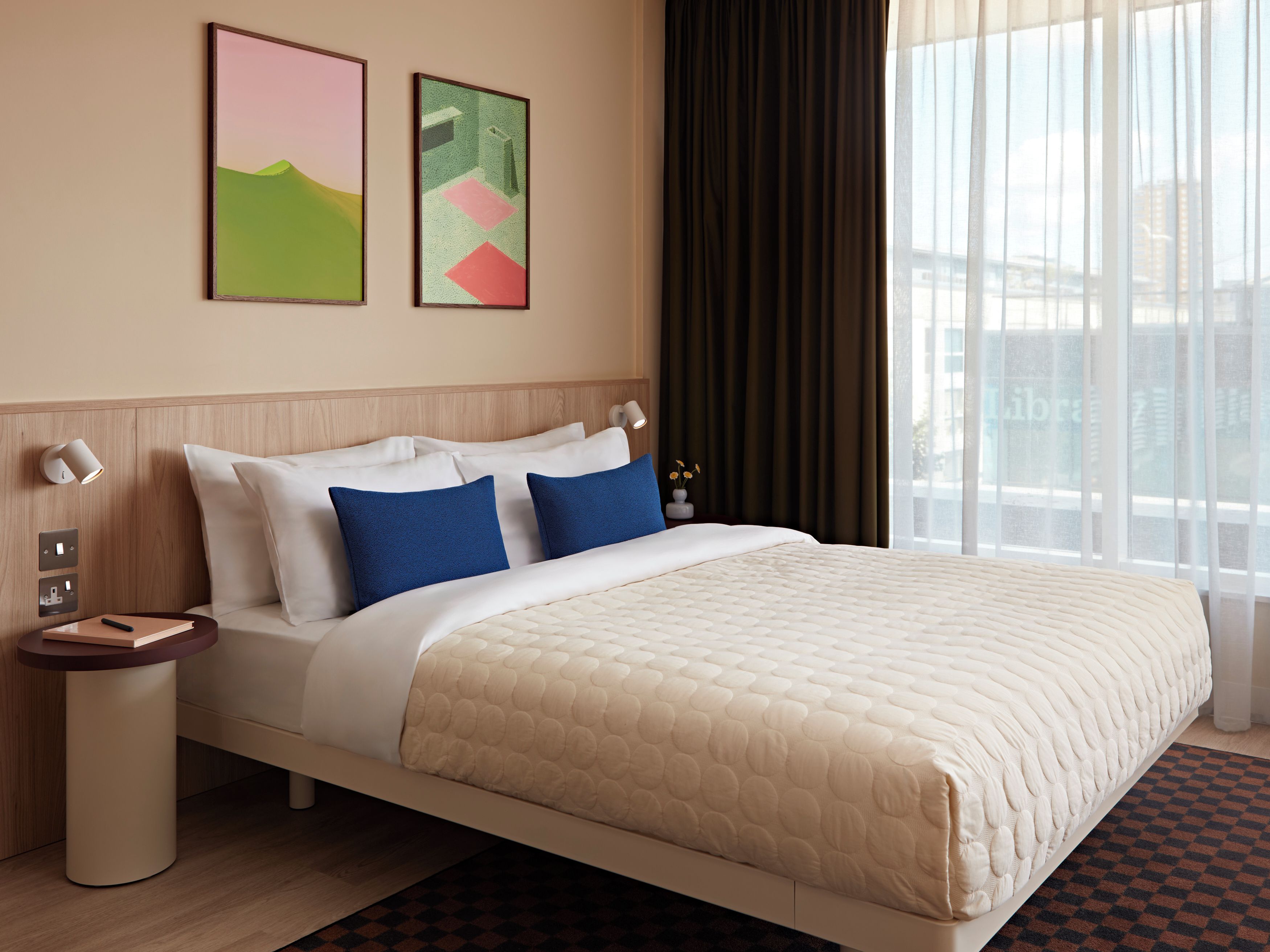 A hotel room with a bed, bedside tables, artwork on the wall, and a window with curtains.