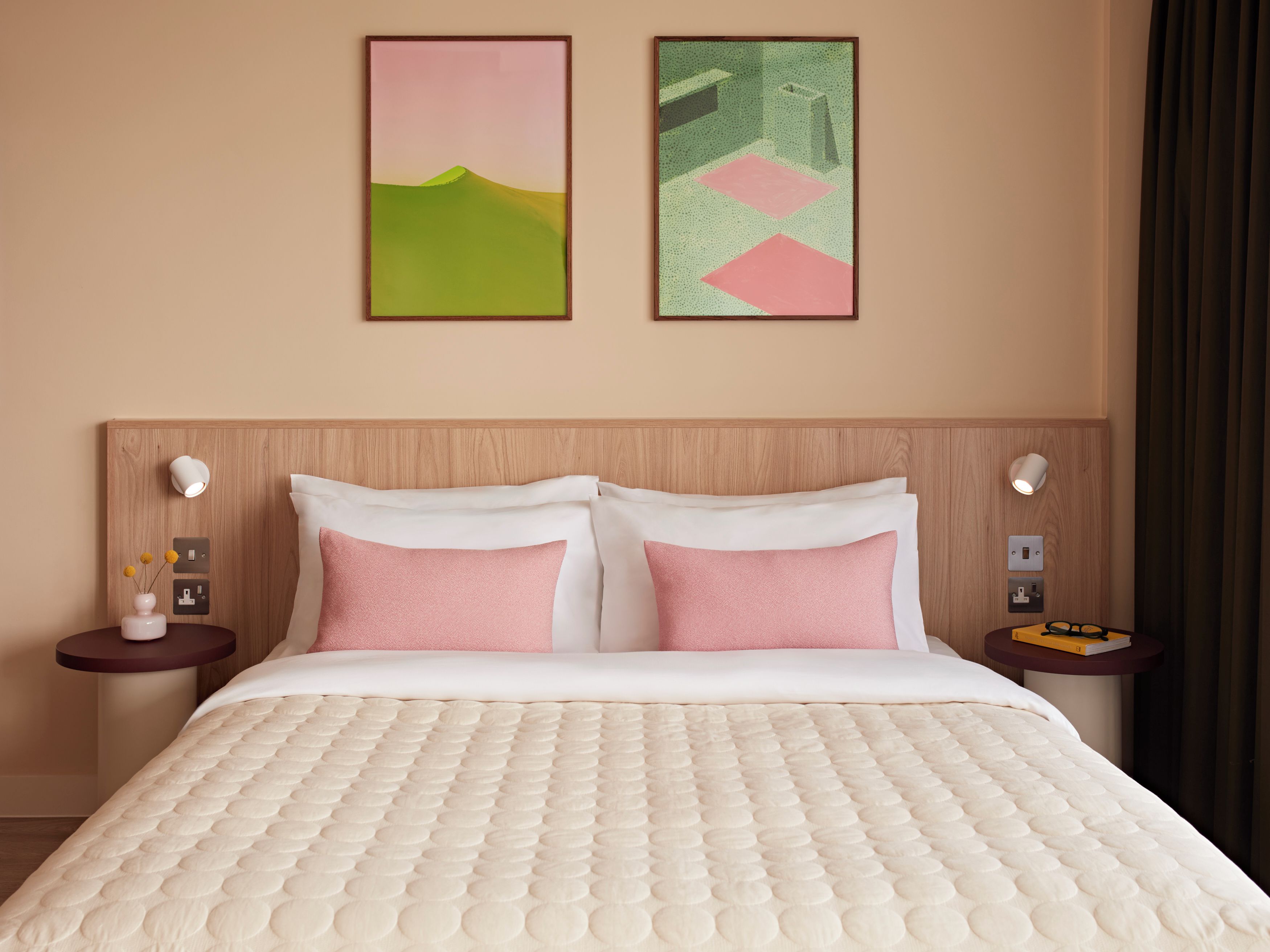 A hotel room with a bed, two bedside tables, a nightstand, a wall-mounted light, and two framed artworks.