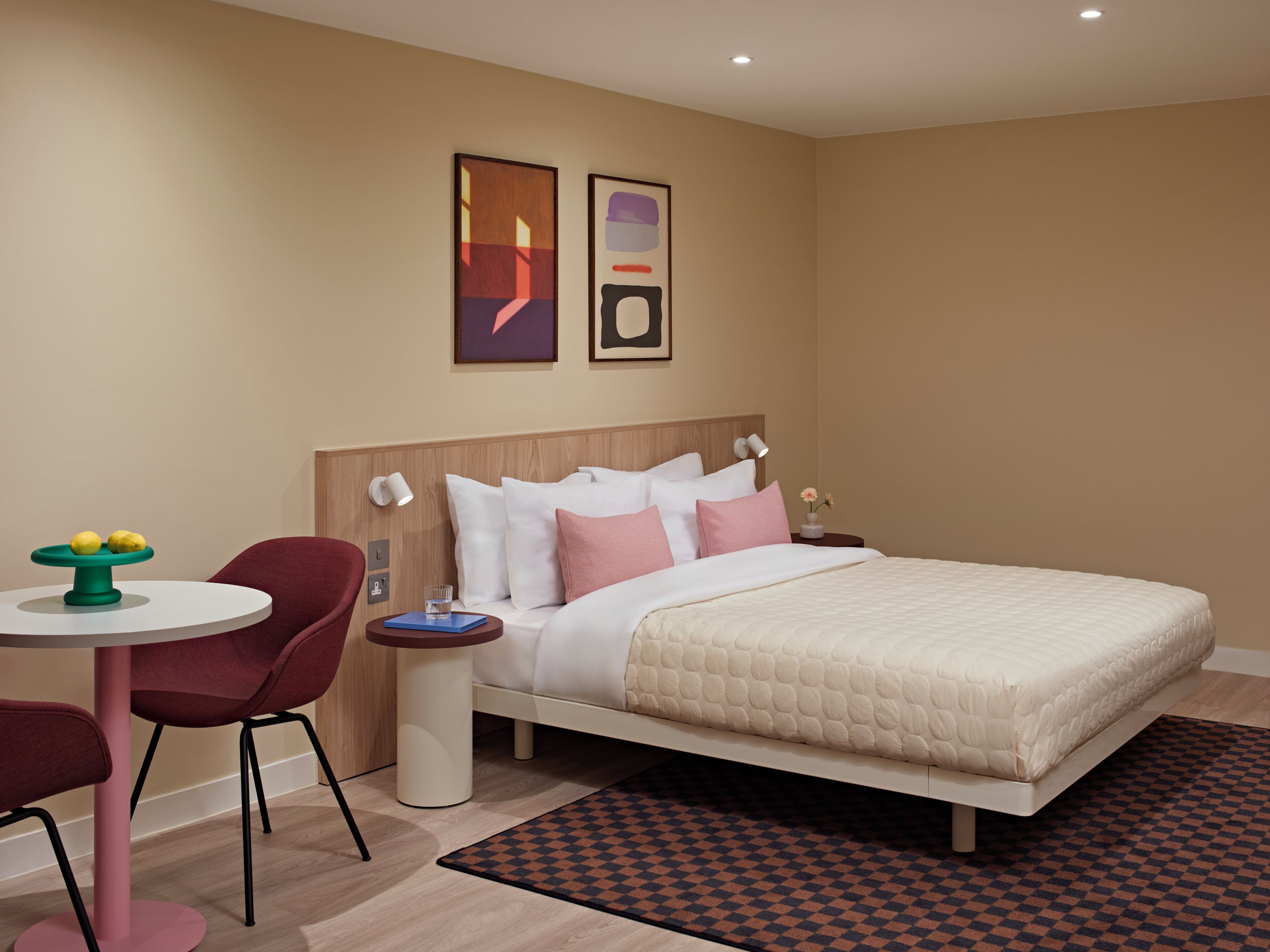 Una habitación de hotel con una cama, un escritorio con una silla, una pequeña mesa con fruta y obras de arte en la pared.