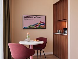 Un comedor moderno con una mesa redonda, dos sillas, una pequeña zona de cocina y un cartel de bienvenida colgado en la pared.