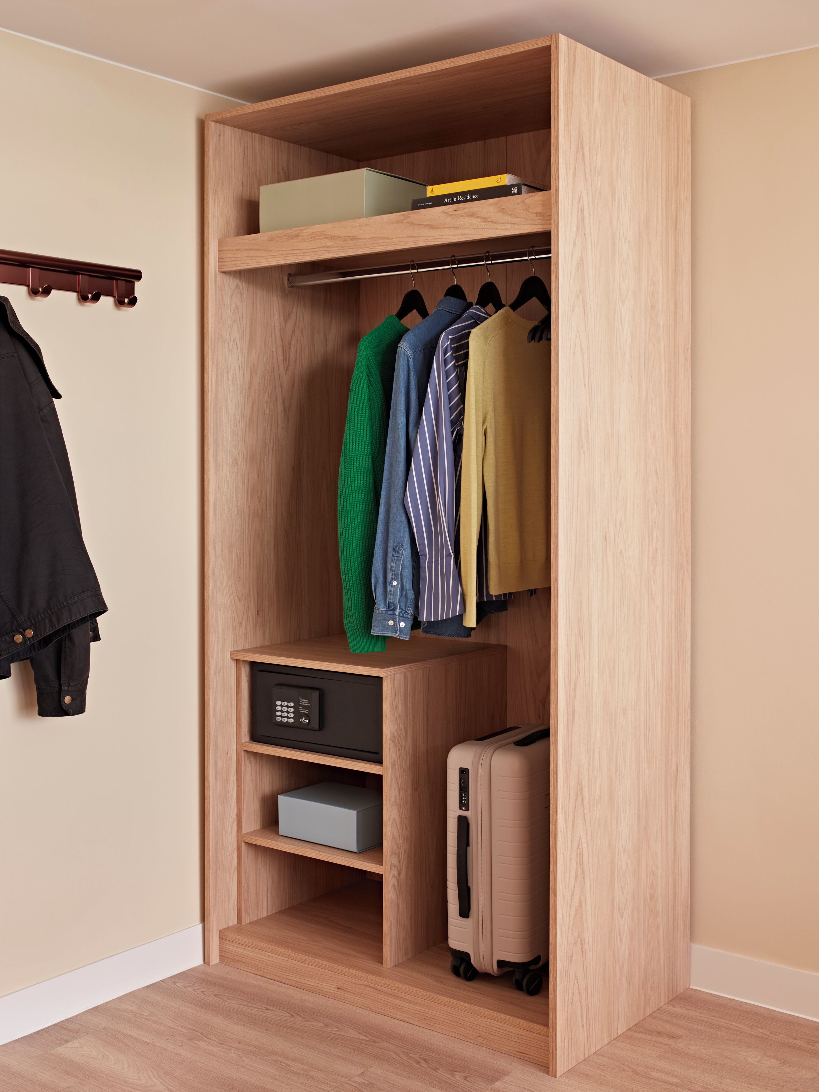 A wooden wardrobe with hanging clothes, a small shelf with books, and a suitcase.