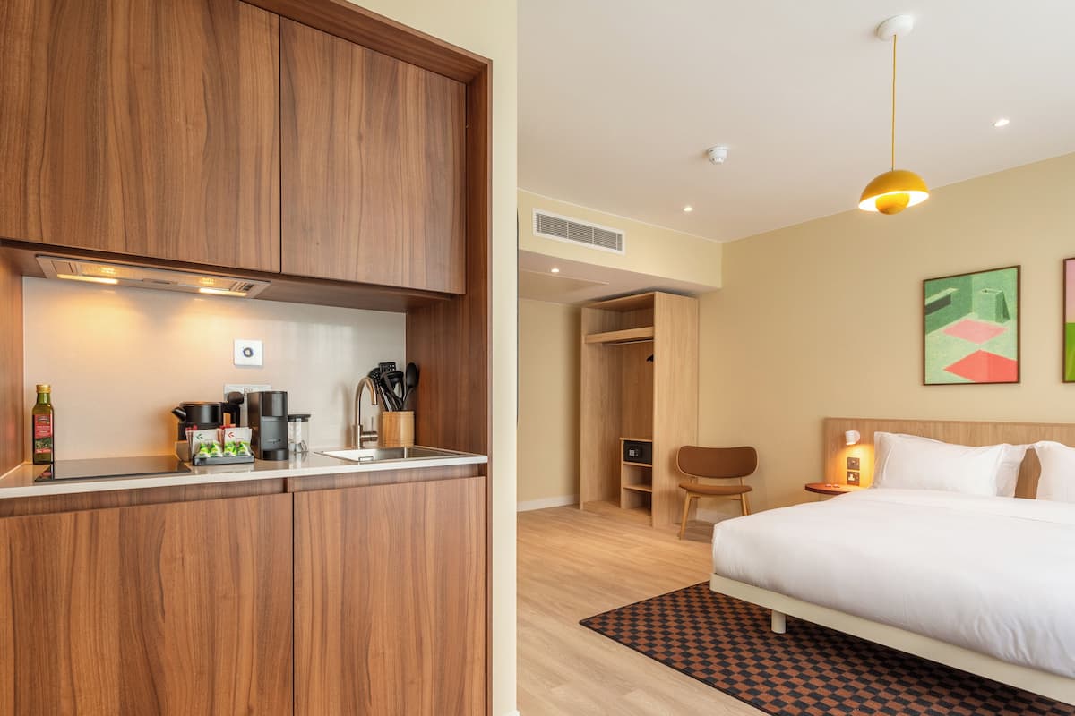 A hotel room with a wooden wardrobe, a kitchenette, a bed, and a small seating area.