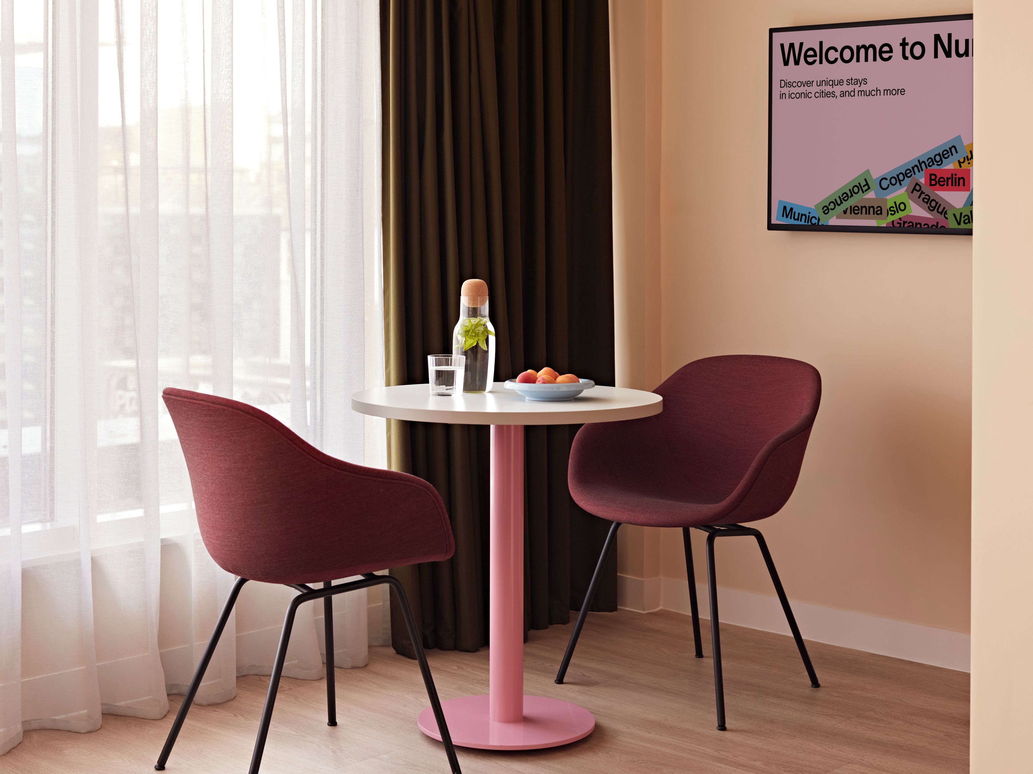 A modern hotel room with a round table, two red chairs, a bottle of water, a glass, and a bowl of fruit.