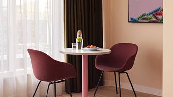 A modern hotel room with a round table, two red chairs, a bottle of water, a glass, and a bowl of fruit.