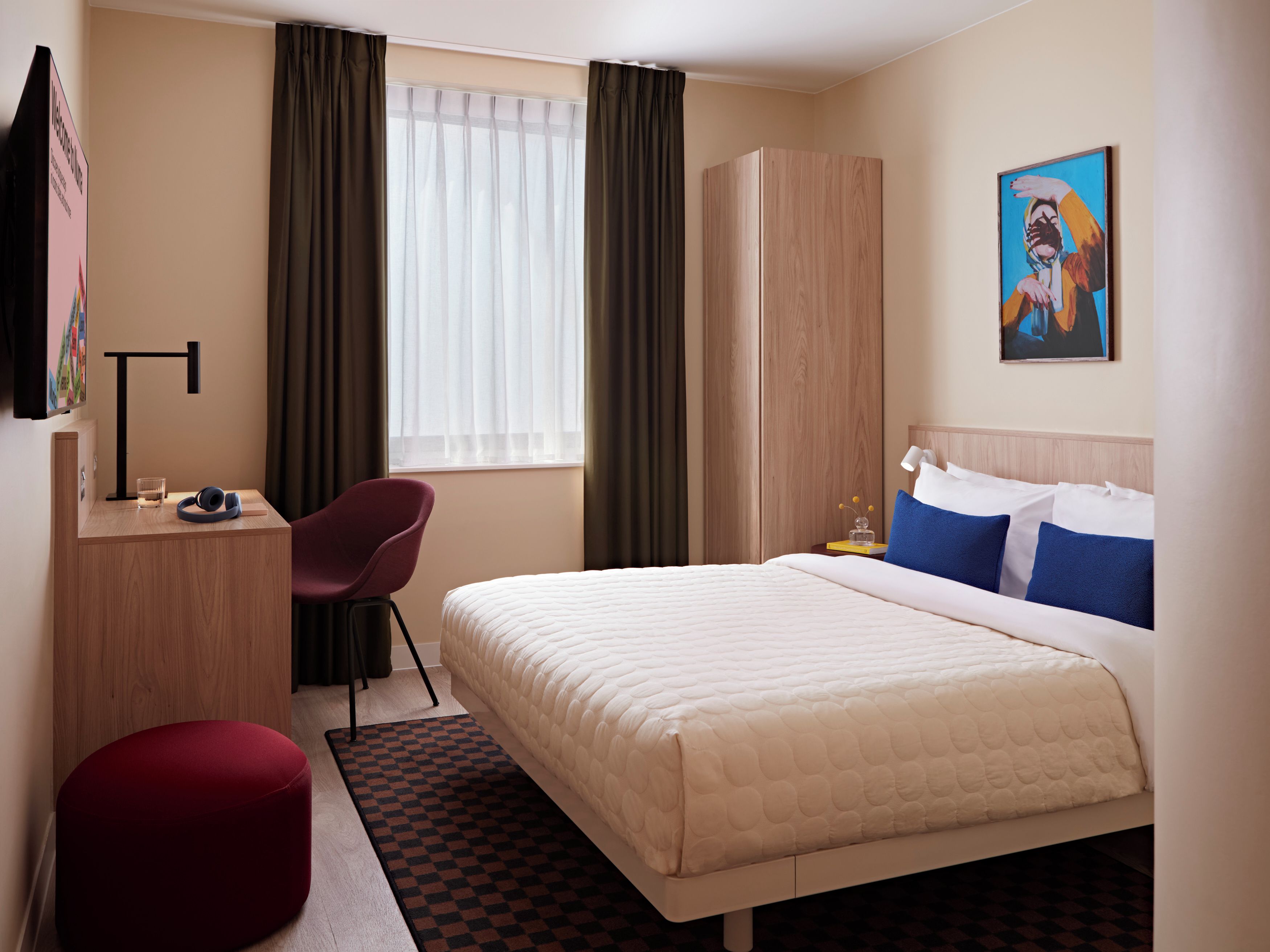 Une chambre d’hôtel avec un lit, un bureau, une chaise, un pouf rouge, un tableau et une fenêtre avec des rideaux.
