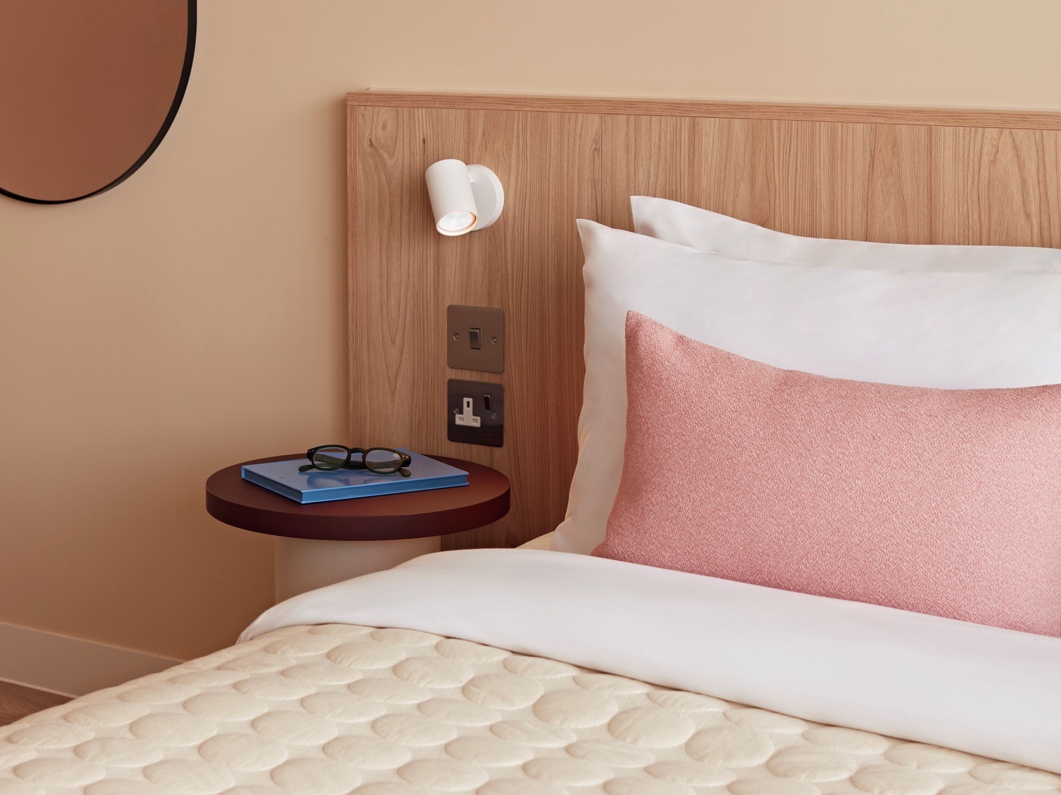A hotel room with a bed, a bedside table with glasses and a notebook, a wall-mounted light, and a power outlet.