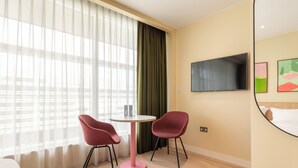 A hotel room with a bed, a TV mounted on the wall, two red chairs around a small table, and a window with sheer curtains. - Numa Brighton Pavilion (Brighton)