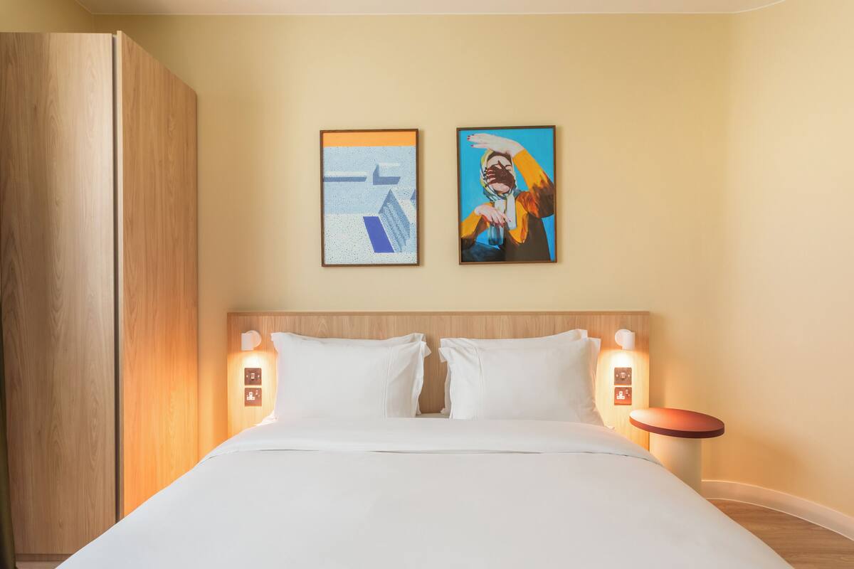 A hotel room with a large bed, two wall-mounted light fixtures, and two framed artworks above the bed.