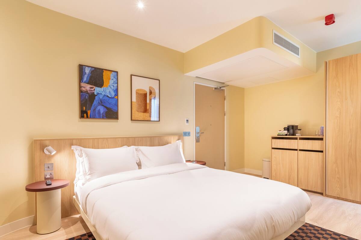 A hotel room with a large bed, a bedside table with a lamp, a small side table with a remote control, a wooden wardrobe, and two framed artworks on the wall.
