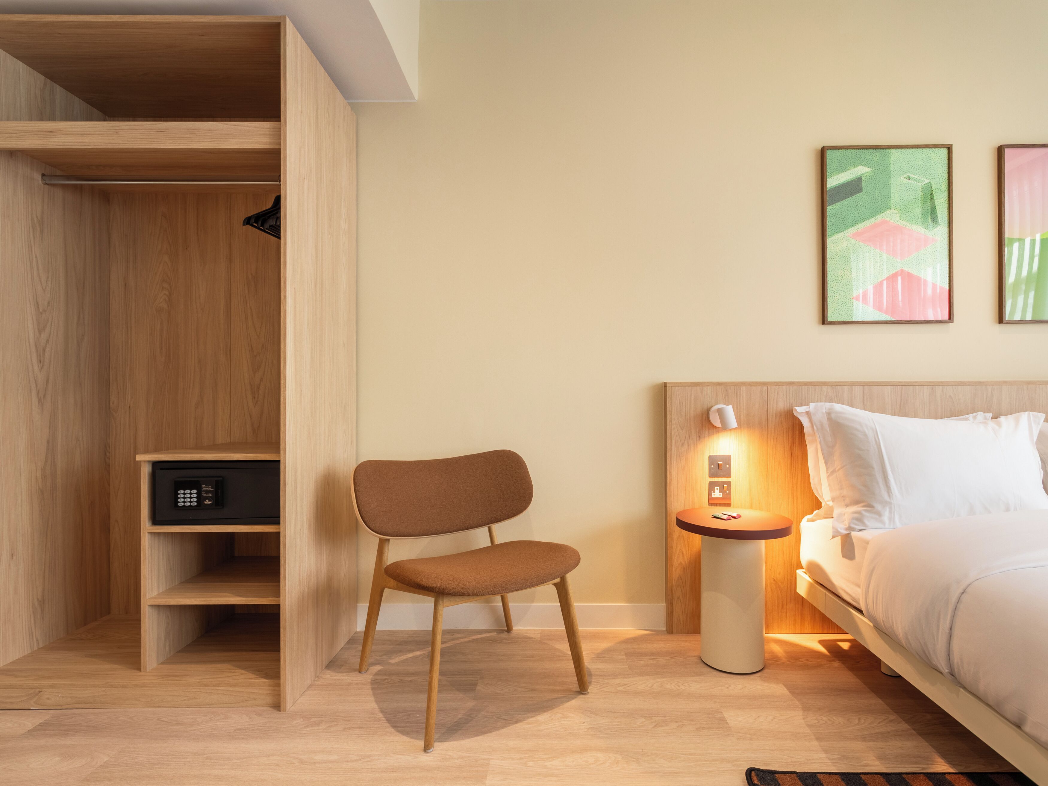 Ein modernes Hotelzimmer mit Bett, Sessel, Nachttisch mit Lampe, Kleiderschrank und Bildern an der Wand.