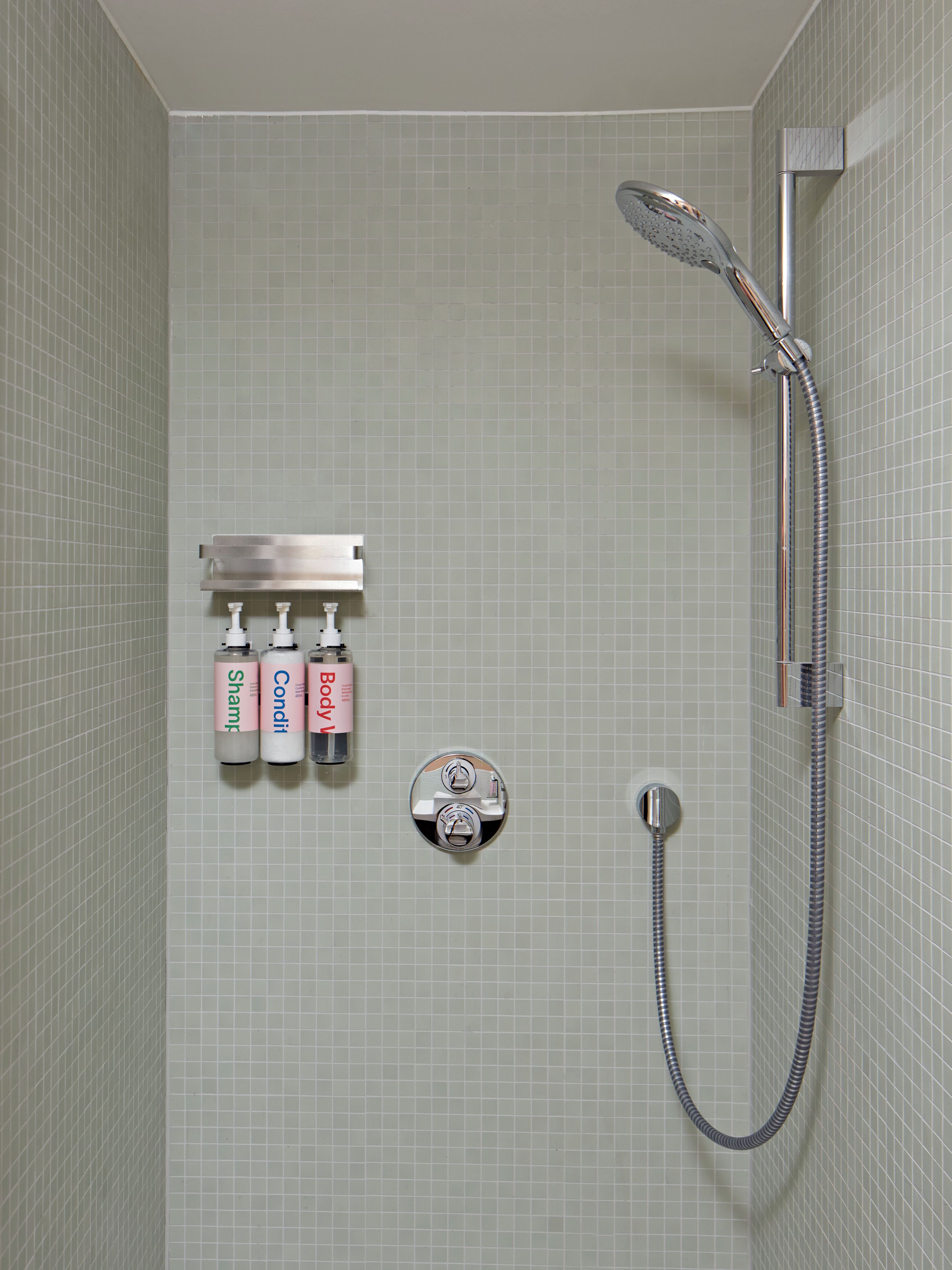 A shower with a handheld showerhead and a wall-mounted faucet. There are three bottles of shampoo, conditioner, and body wash on a shelf.