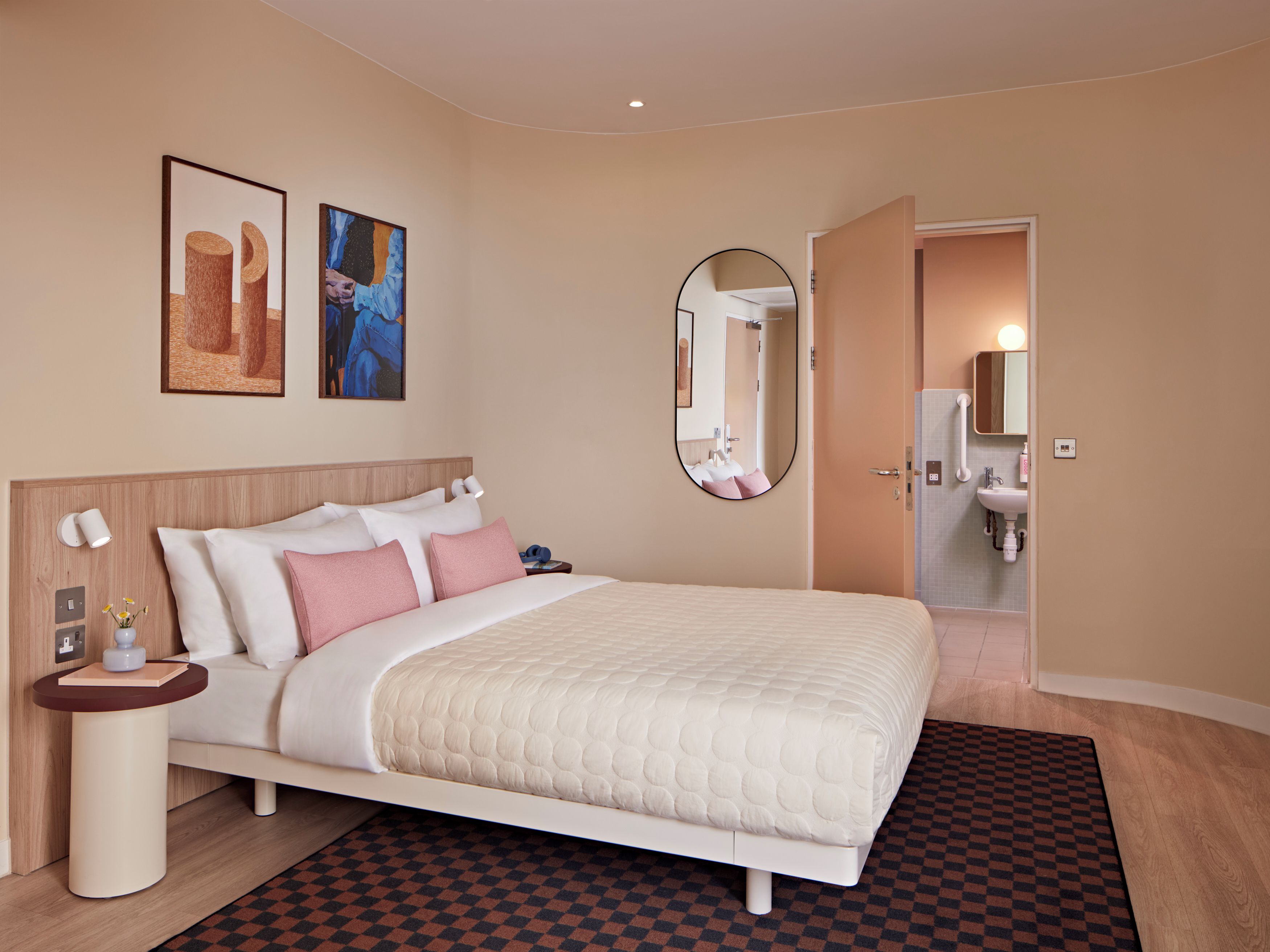 Une chambre d’hôtel avec un lit, une table de chevet, des œuvres d’art mural, un miroir et une salle de bain visible à travers une porte ouverte.