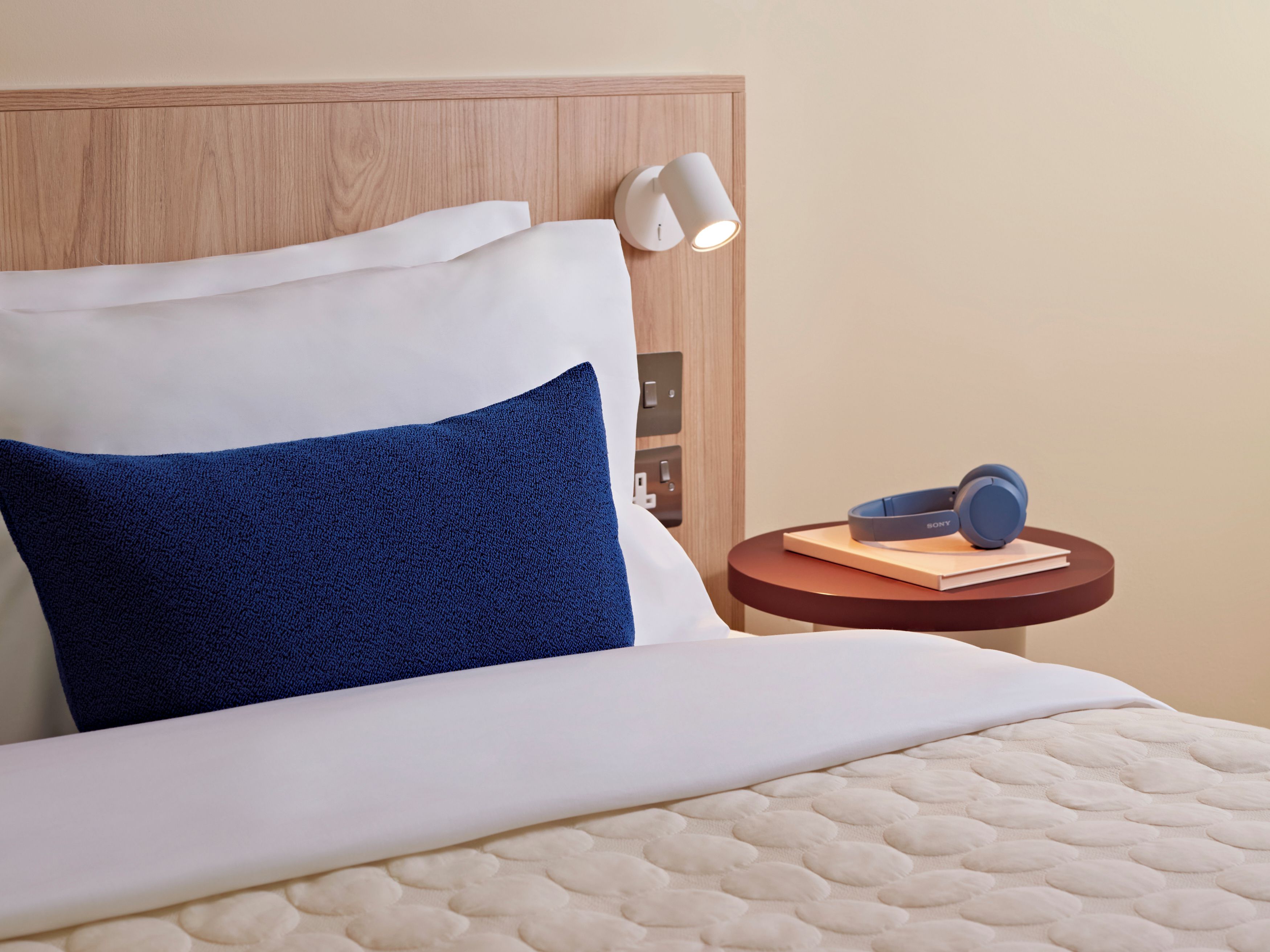 A bed with white and blue pillows, a round bedside table with a book and headphones, and a wall-mounted lamp.