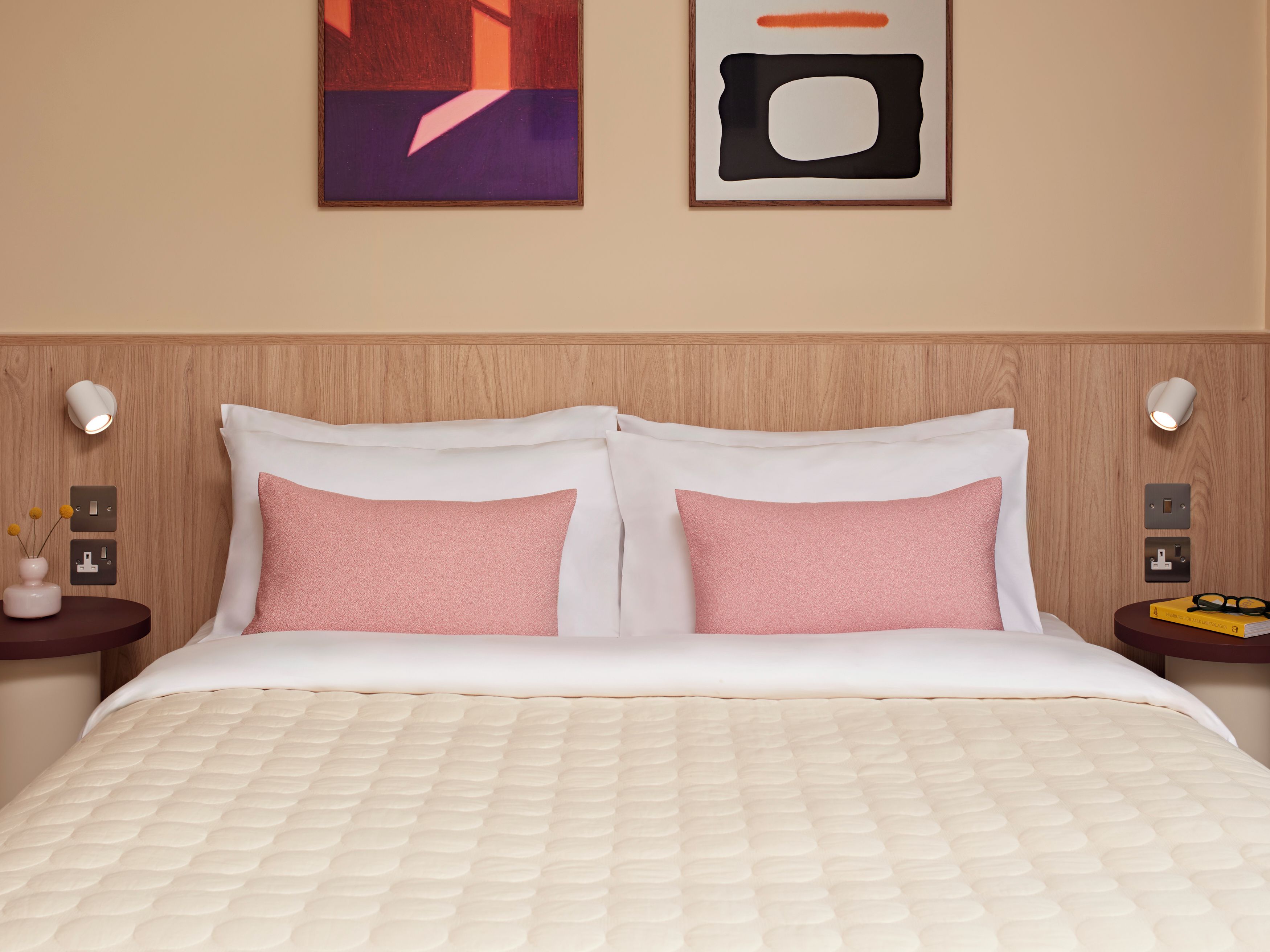 A hotel room with a bed, two bedside tables, a wall-mounted light fixture, and a framed abstract painting.