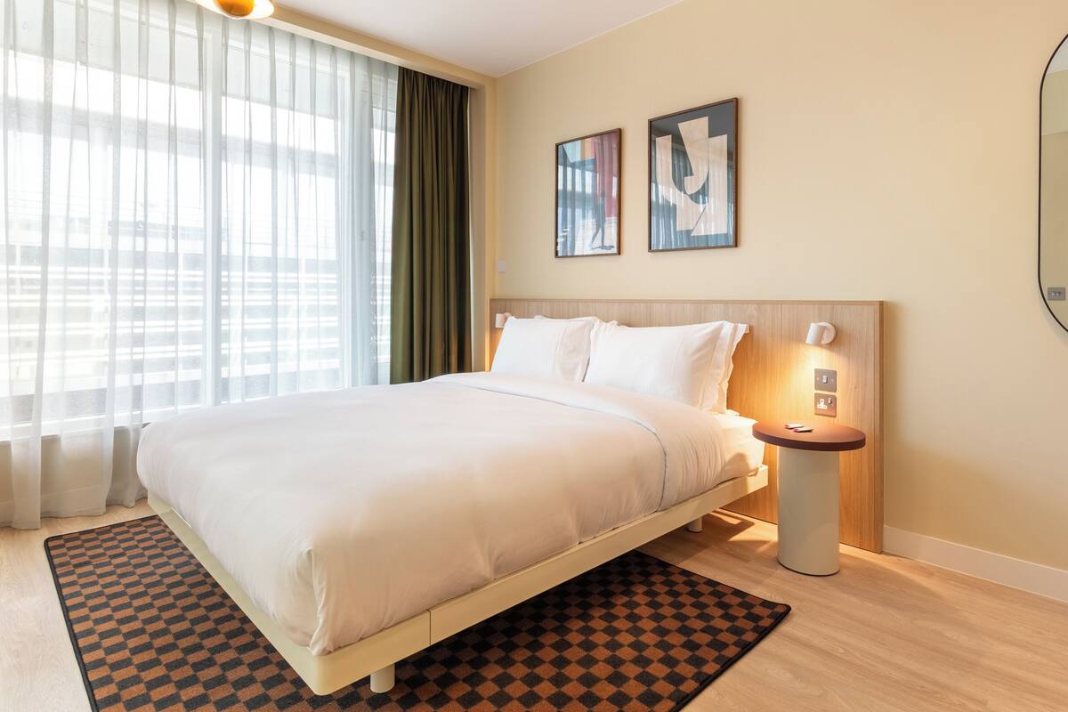 A hotel room with a large bed, a bedside table, a nightstand, a lamp, and two framed artworks on the wall.