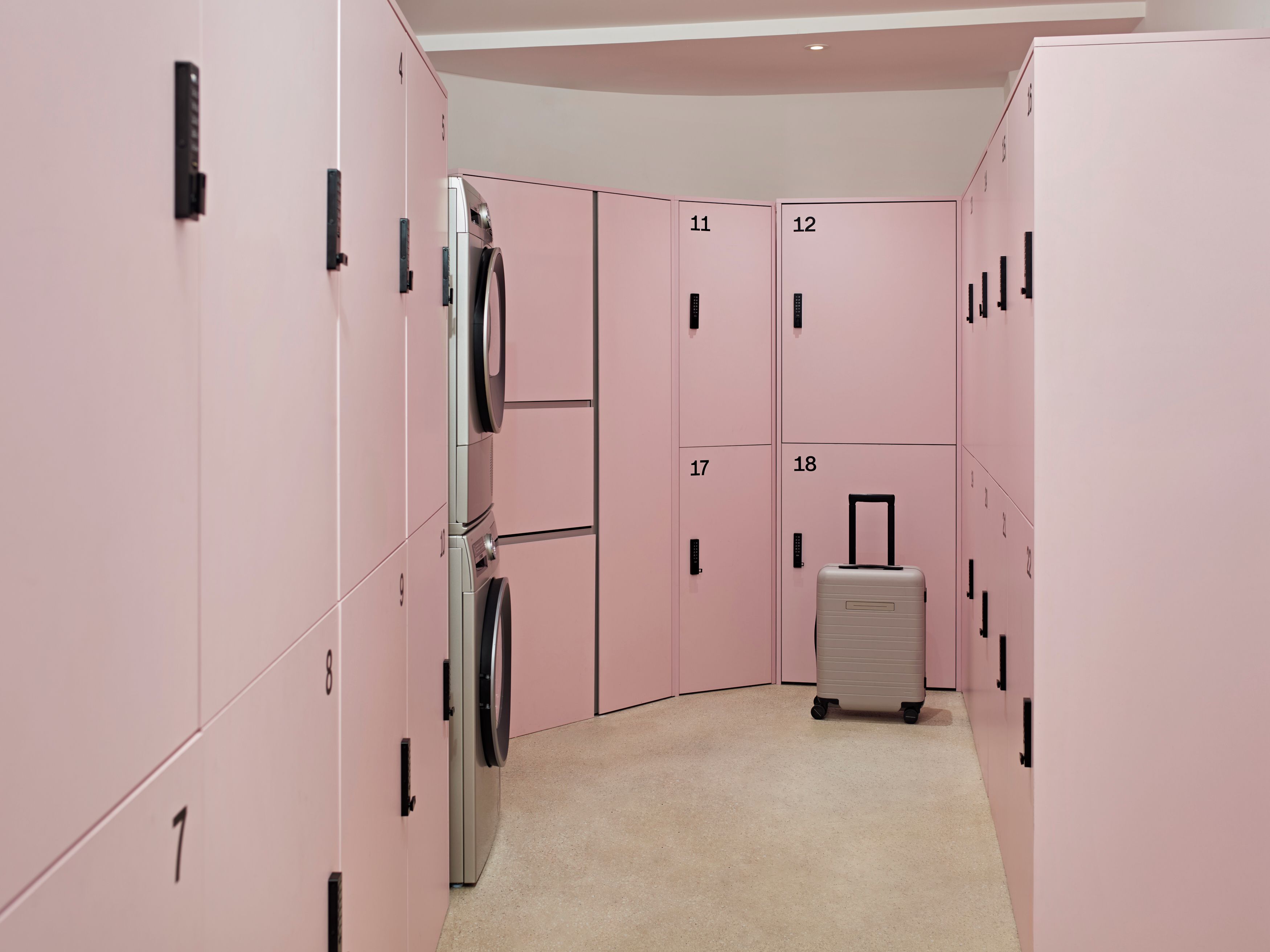 A corridor with pink lockers numbered 1 to 18, a washing machine, and a suitcase.