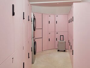 Un pasillo con taquillas rosas numeradas del 1 al 18, una lavadora y una maleta.