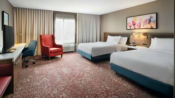 A hotel room with two beds, a desk with a chair, a red armchair, a TV, and a large window with curtains.