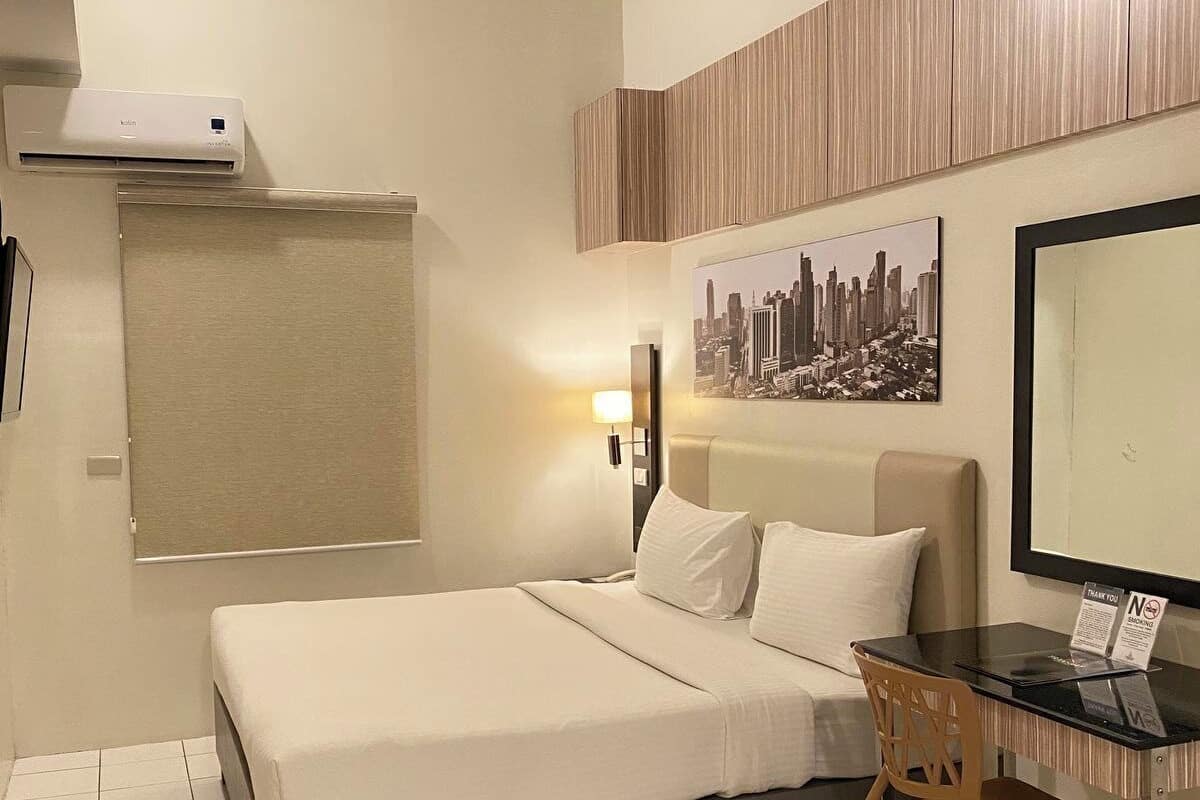 A hotel room with a bed, a desk, a chair, a cityscape painting, and an air conditioning unit.