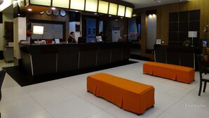 Un espace d’accueil d’hôtel comprenant un comptoir de réception, deux coins salon avec des sièges rembourrés de couleur orange et une horloge murale.