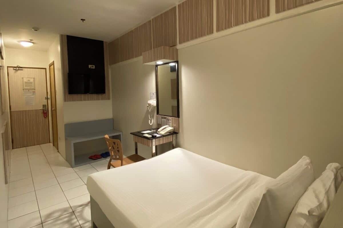 A hotel room with a large bed, a desk with a telephone, a mirror, and a door leading to a hallway.