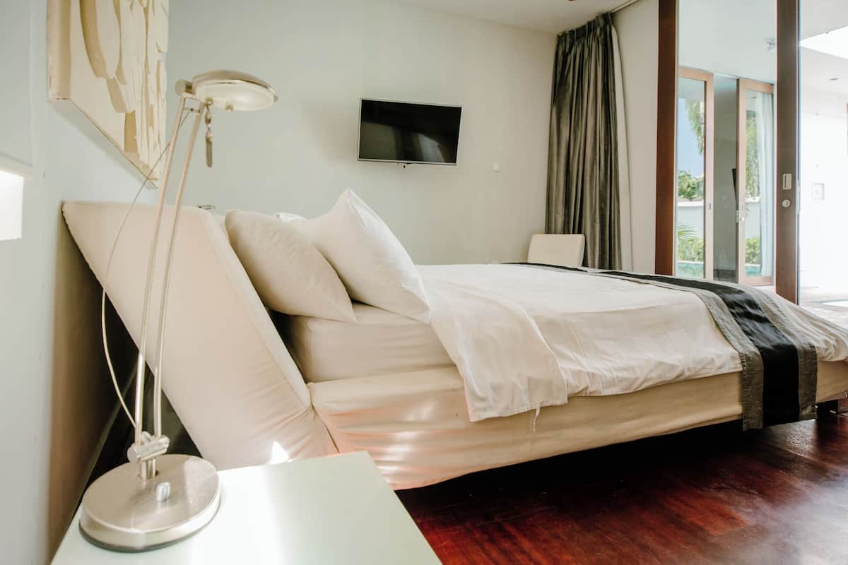 Uma cama bem arrumada com edredom branco e travesseiros, uma luminária de cabeceira e uma TV de tela plana na parede.