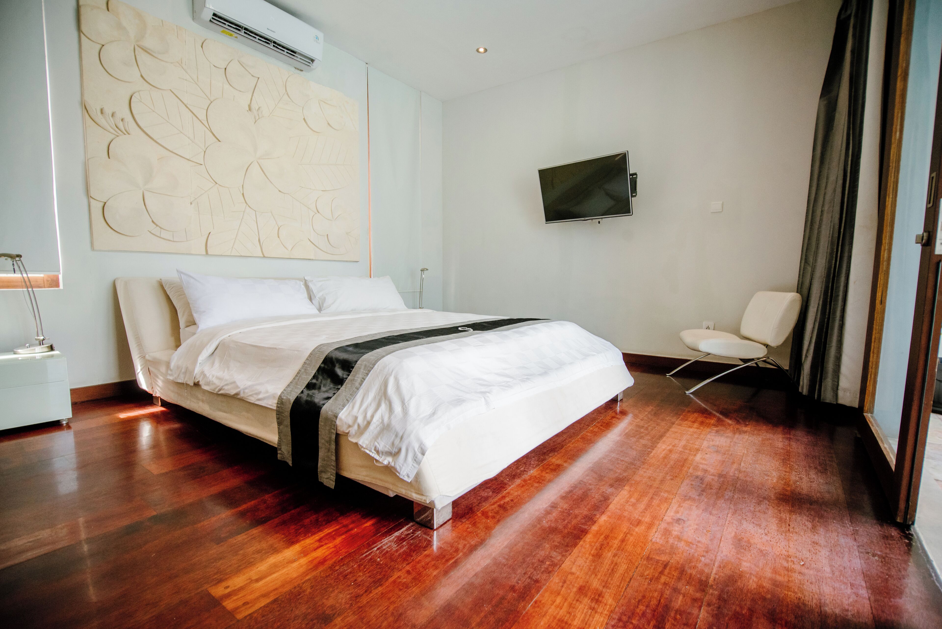 A modern bedroom with a wooden floor, a bed with white bedding, a mounted TV, and a chair.
