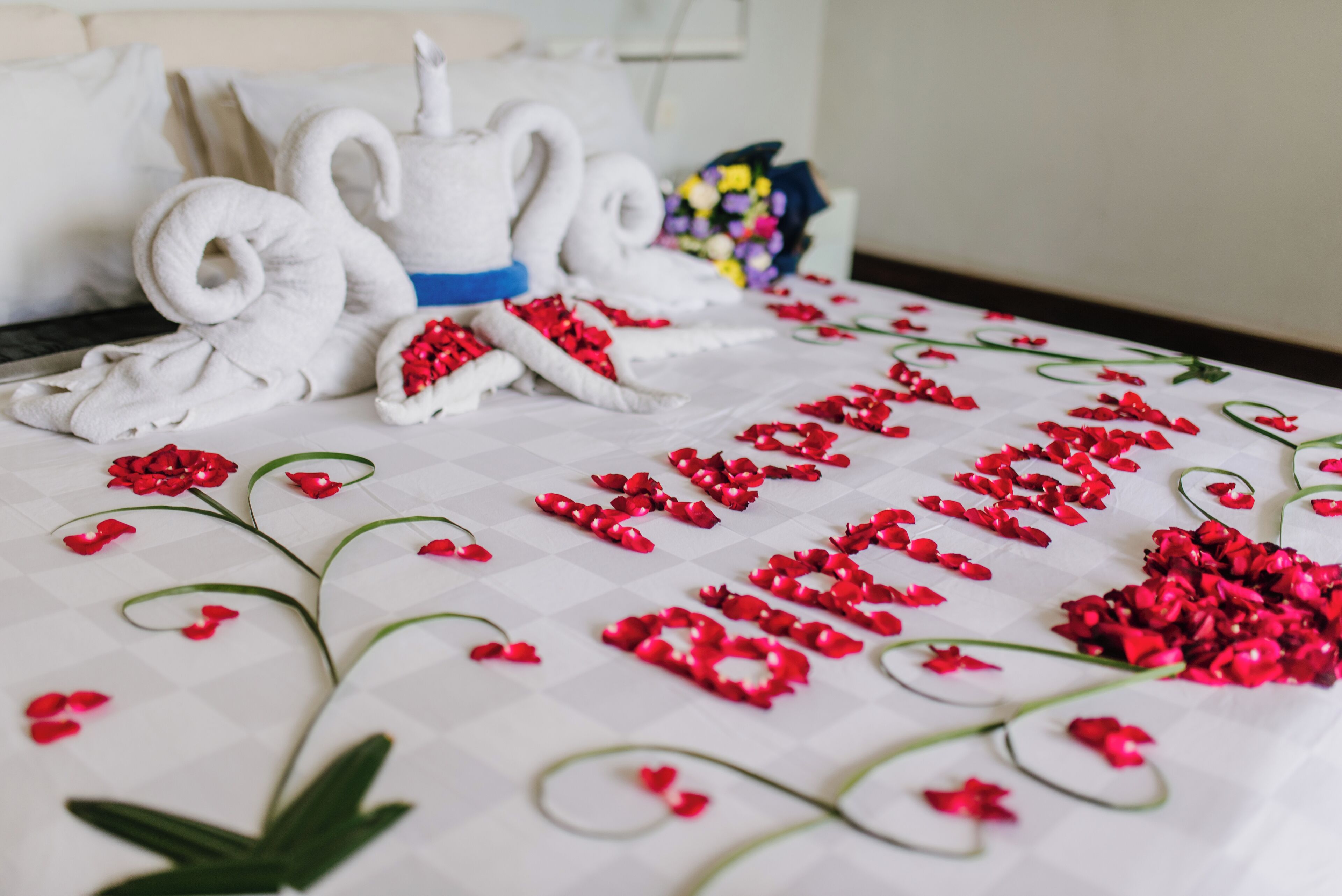A bed with a white bedspread, red rose petals arranged in the shape of "Happy Birthday," and white towels folded into swans.